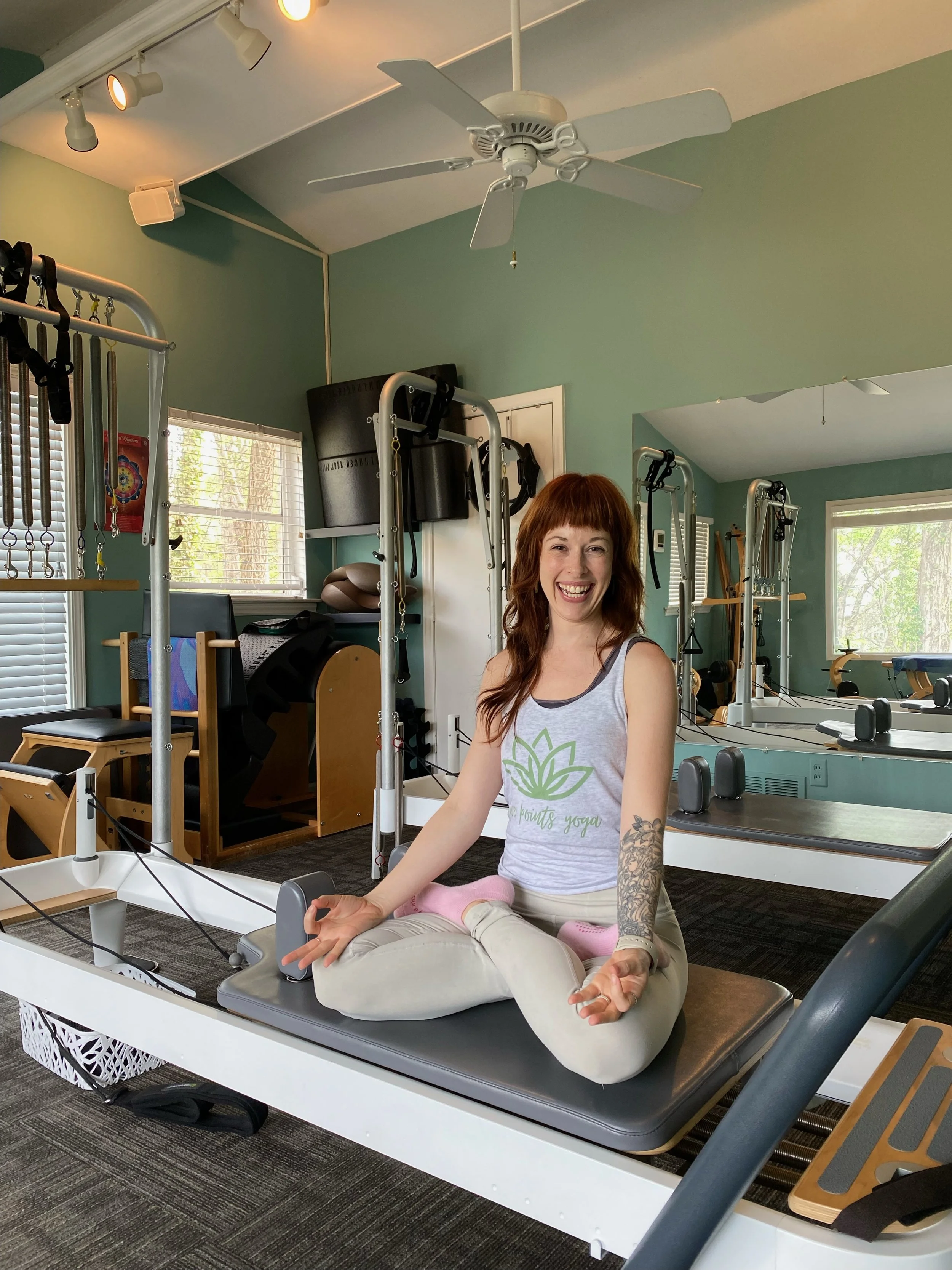 Pilates instructor sitting on reformer carriage in meditative pose while smiling at the camera