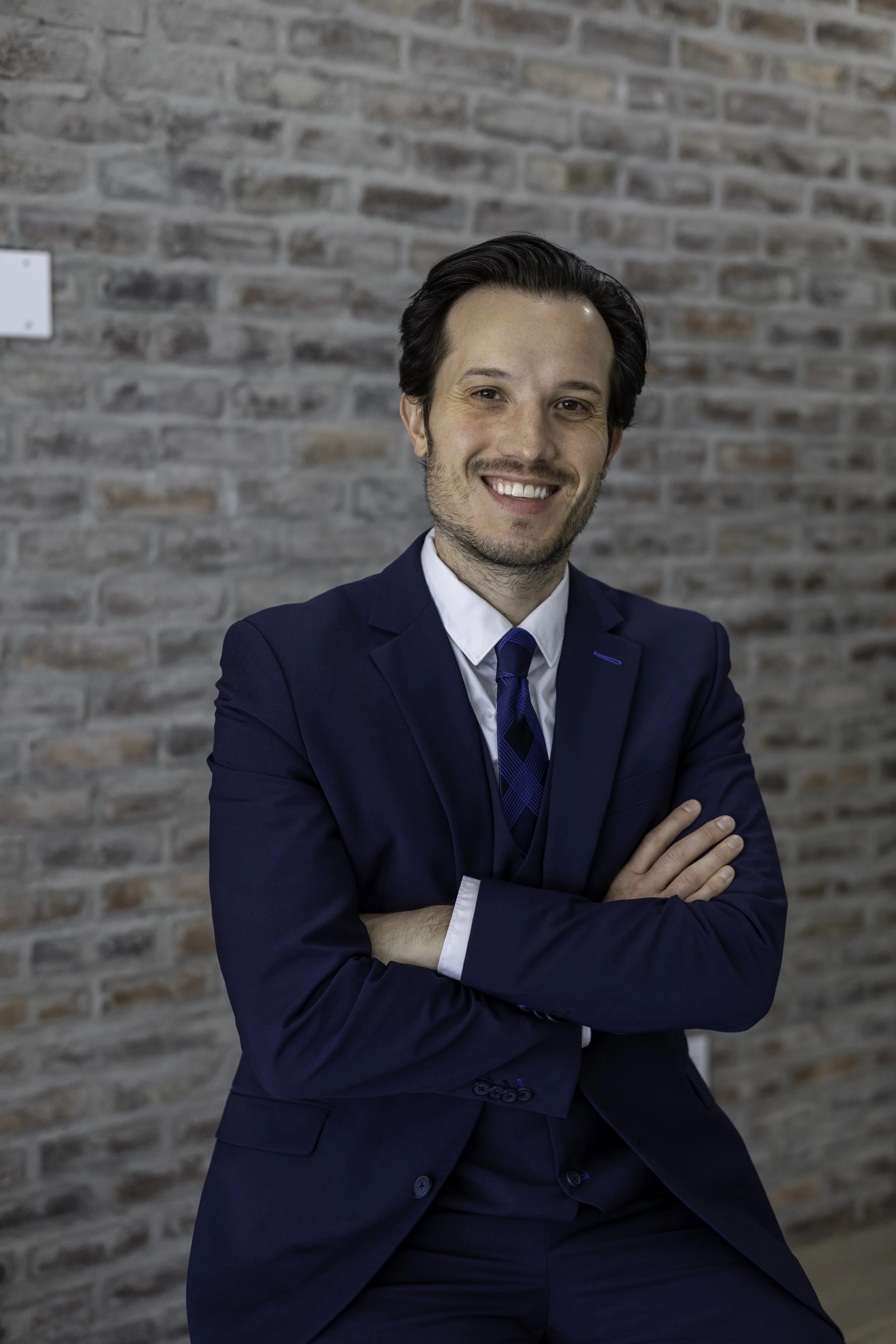 Professional man in a navy suit with arms crossed, standing against a brick wall and smiling.