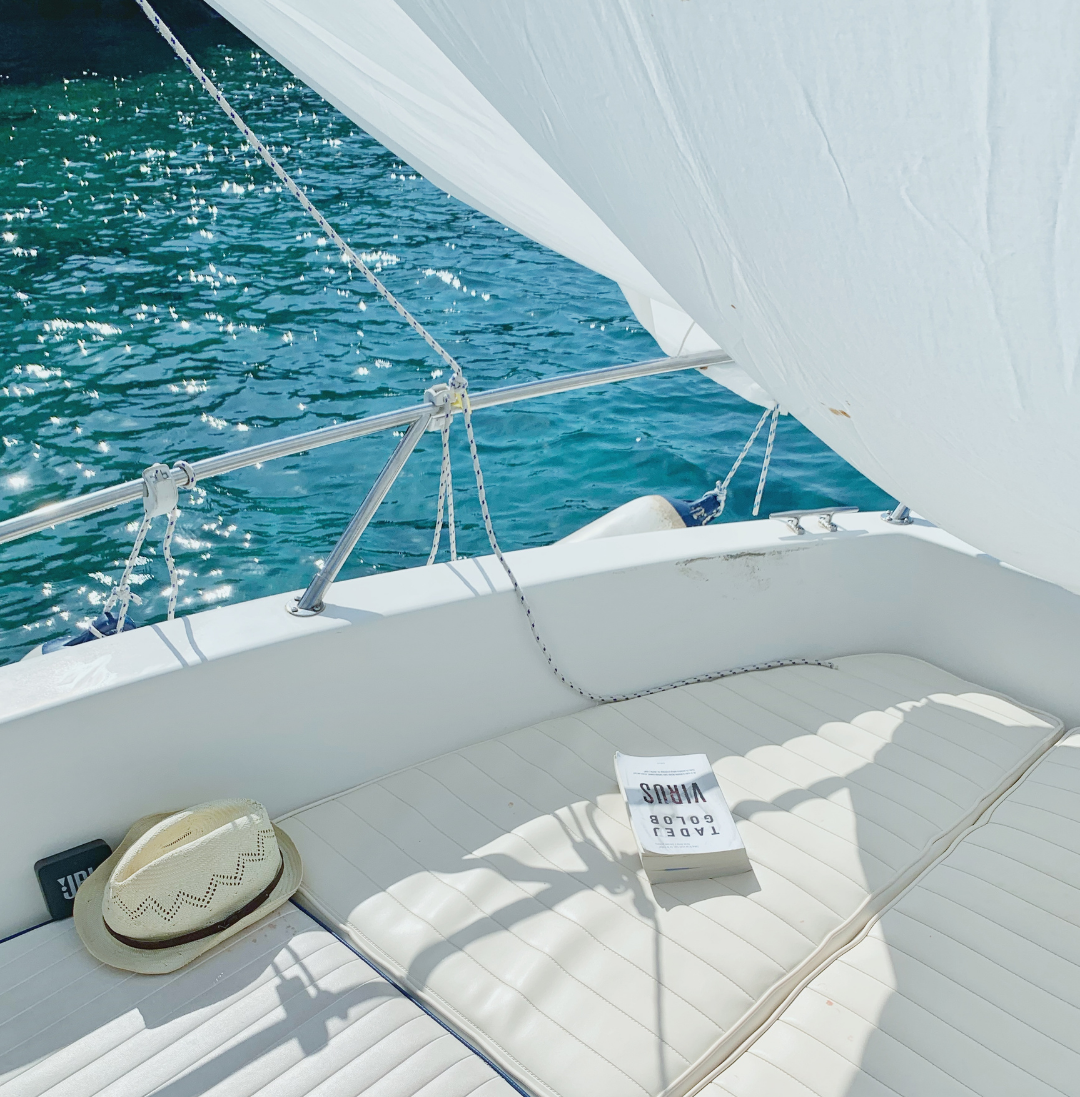 View of a white sailboat deck with a book titled "TADE GOLDBY VIKAS" and a straw hat, with blue water and a sail in the background.