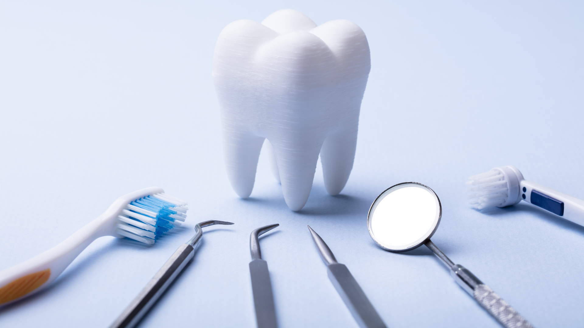 Dental tools and toothbrushes arranged around a large model tooth on a light blue background, representing oral hygiene and preventive dental care.