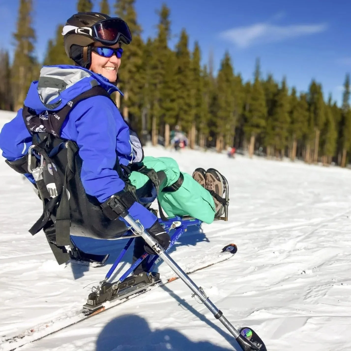 A person in winter gear sitting and smiling on a snow-covered slope with a forest of pine trees in the background, holding ski poles and wearing skis.