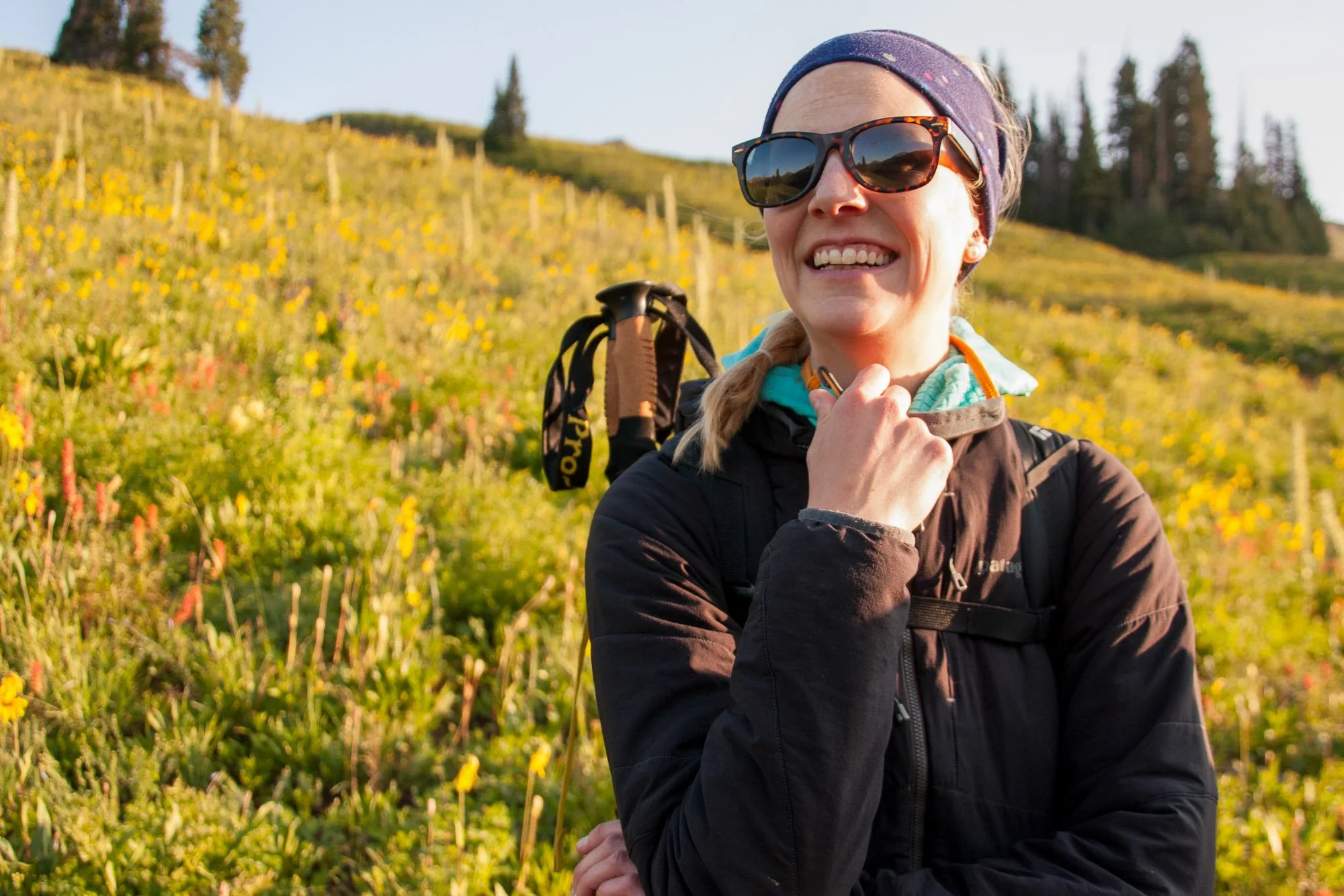 A woman with sunglasses and a headband outdoors in a hillside field of yellow and orange flowers, smiling.