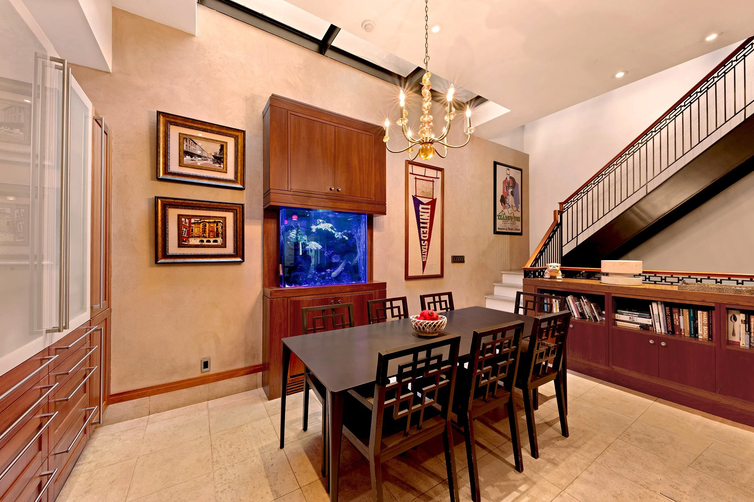A dining room with a black rectangular table and eight matching chairs, a wooden cabinet with a fish tank, framed artwork on the walls, a staircase with a black railing, and a chandelier hanging from the ceiling at 152 Reade Street in Tribeca.
