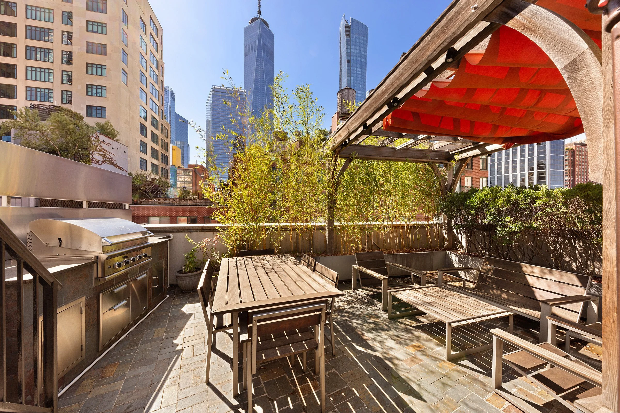 City rooftop patio with outdoor furniture, a barbecue grill, potted plants, bamboo trees, and a view of tall skyscrapers including One World Trade Center under a clear blue sky at 152 Reade Street in Tribeca.