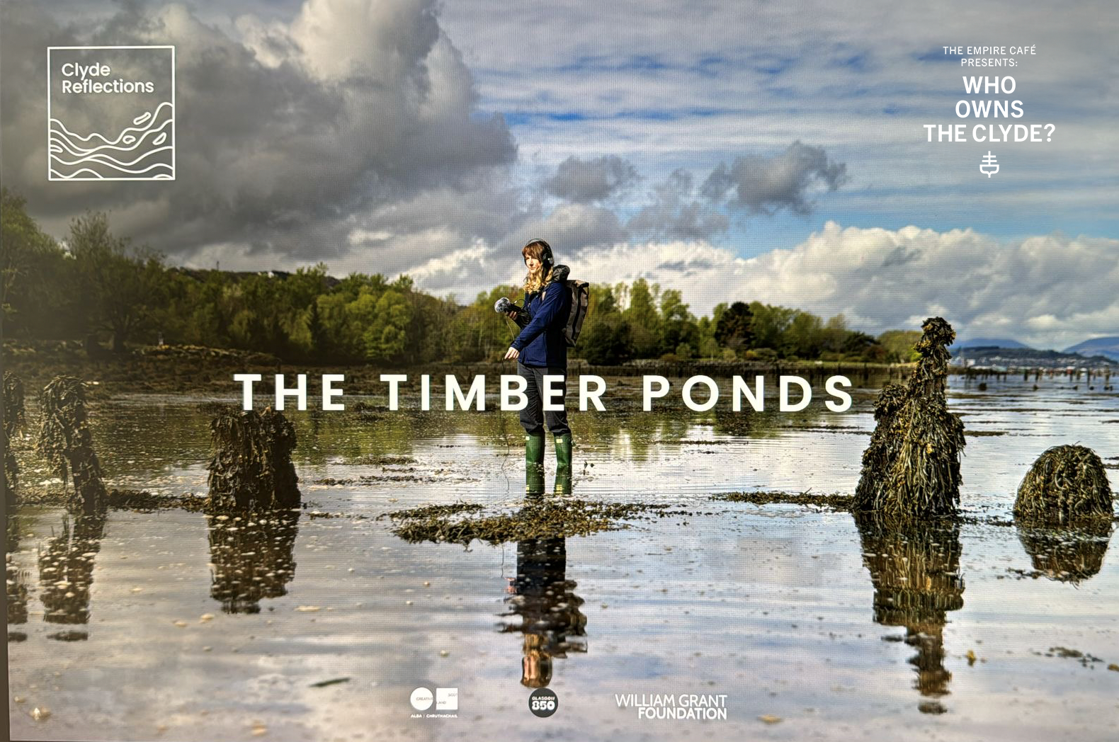 A woman standing in Timber Ponds, a tidal wetland with wooden posts and water reflections, holding binoculars, with cloudy sky and green trees in the background. Text overlays include "The Timber Ponds" and "The Empire Café Presents: Who Owns the Clyde?" along with logos and the William Grant Foundation.