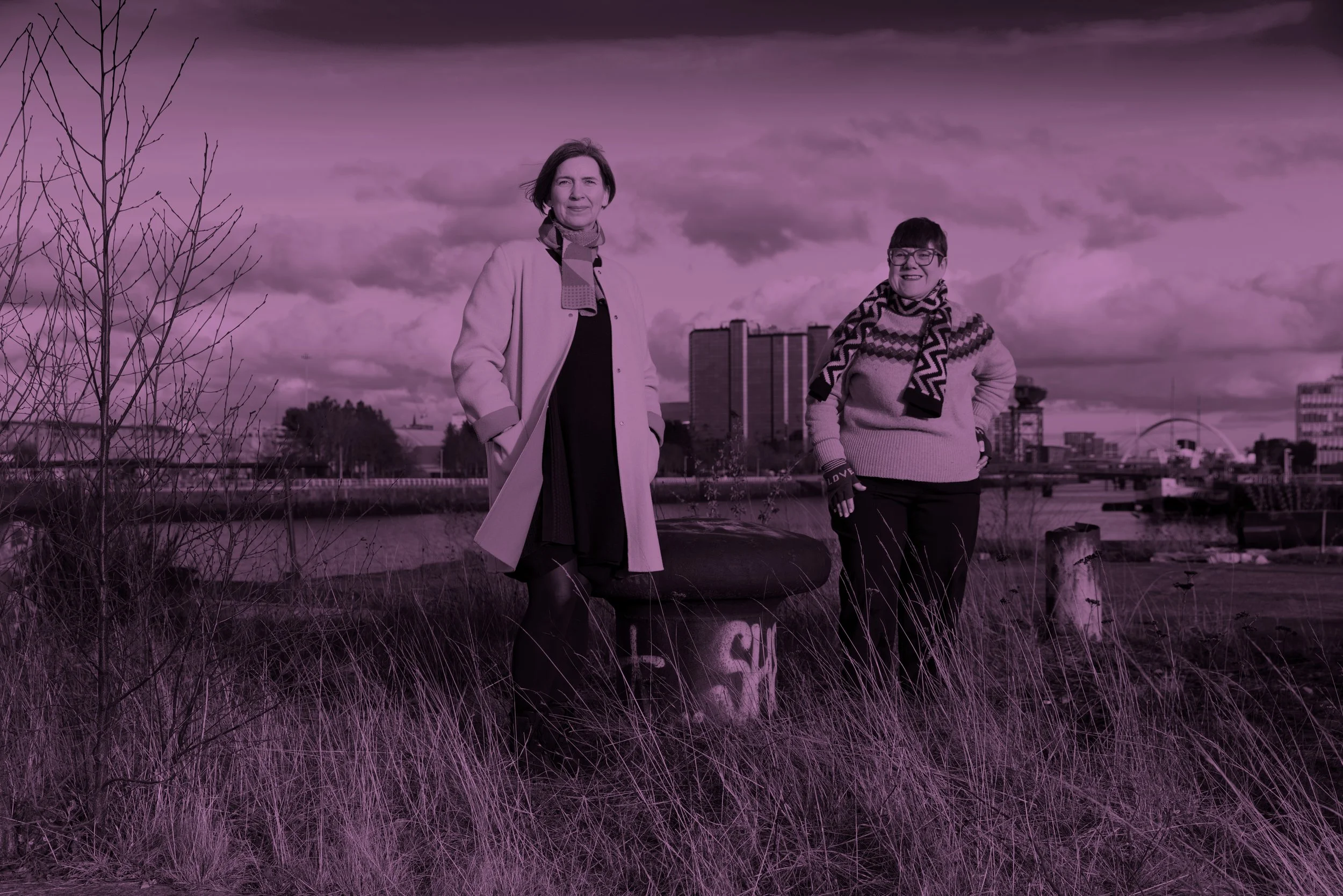 Two women standing outdoors in a grassy area with tall grass and a city skyline in the background, under a cloudy sky.