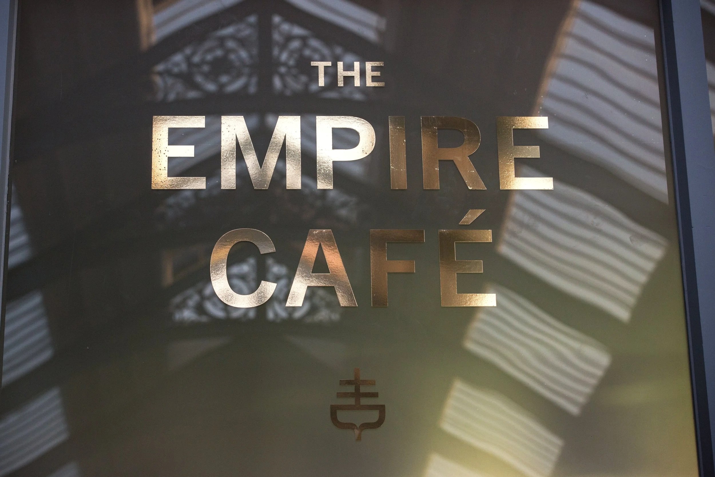 Sign for The Empire Café with metallic text and a decorative emblem below, reflecting nearby striped awnings and architectural details.