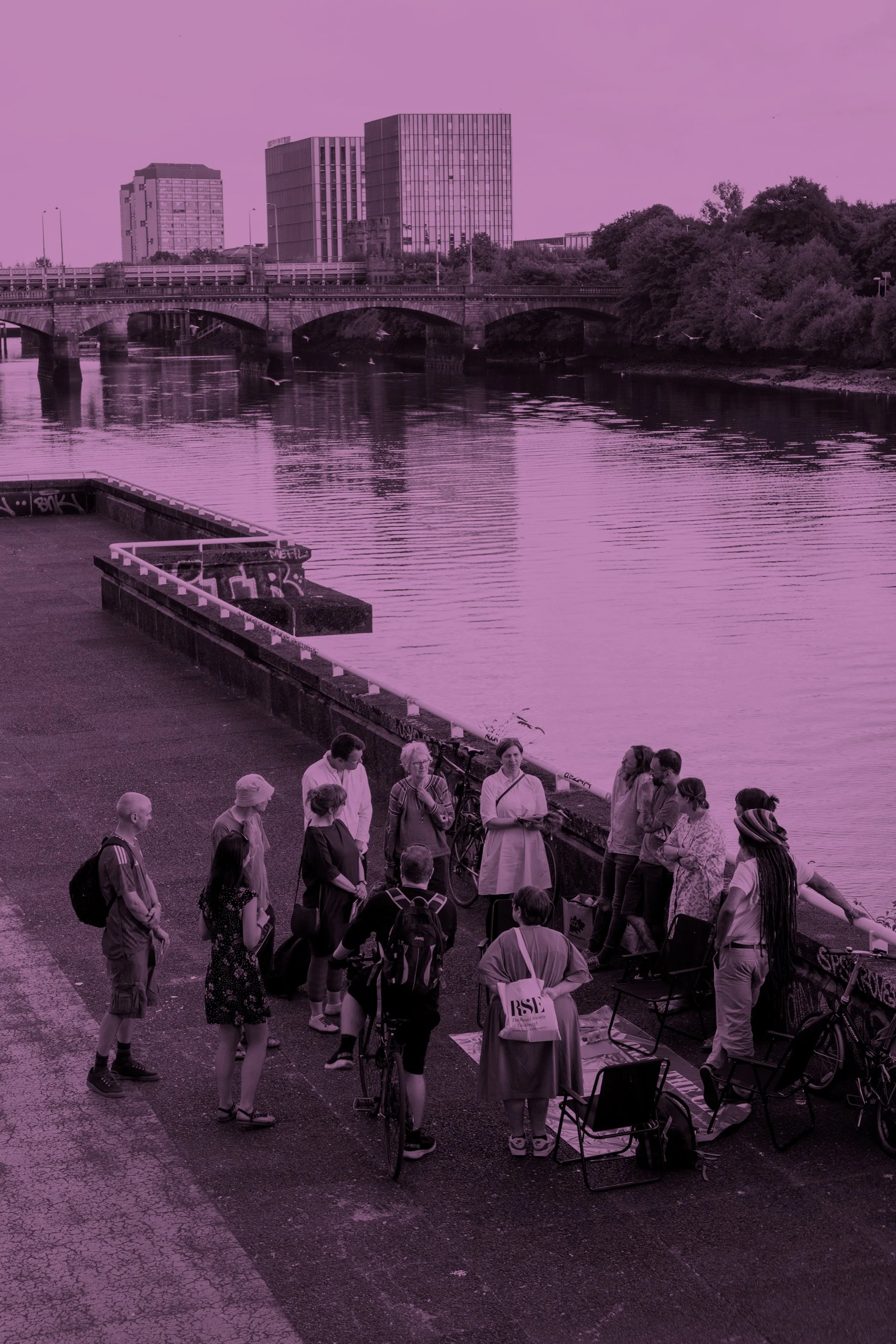 A group of people gathered on a riverside promenade with a bridge in the background, overlooking the water with office buildings and trees on the far bank, during daylight with a purple overlay.