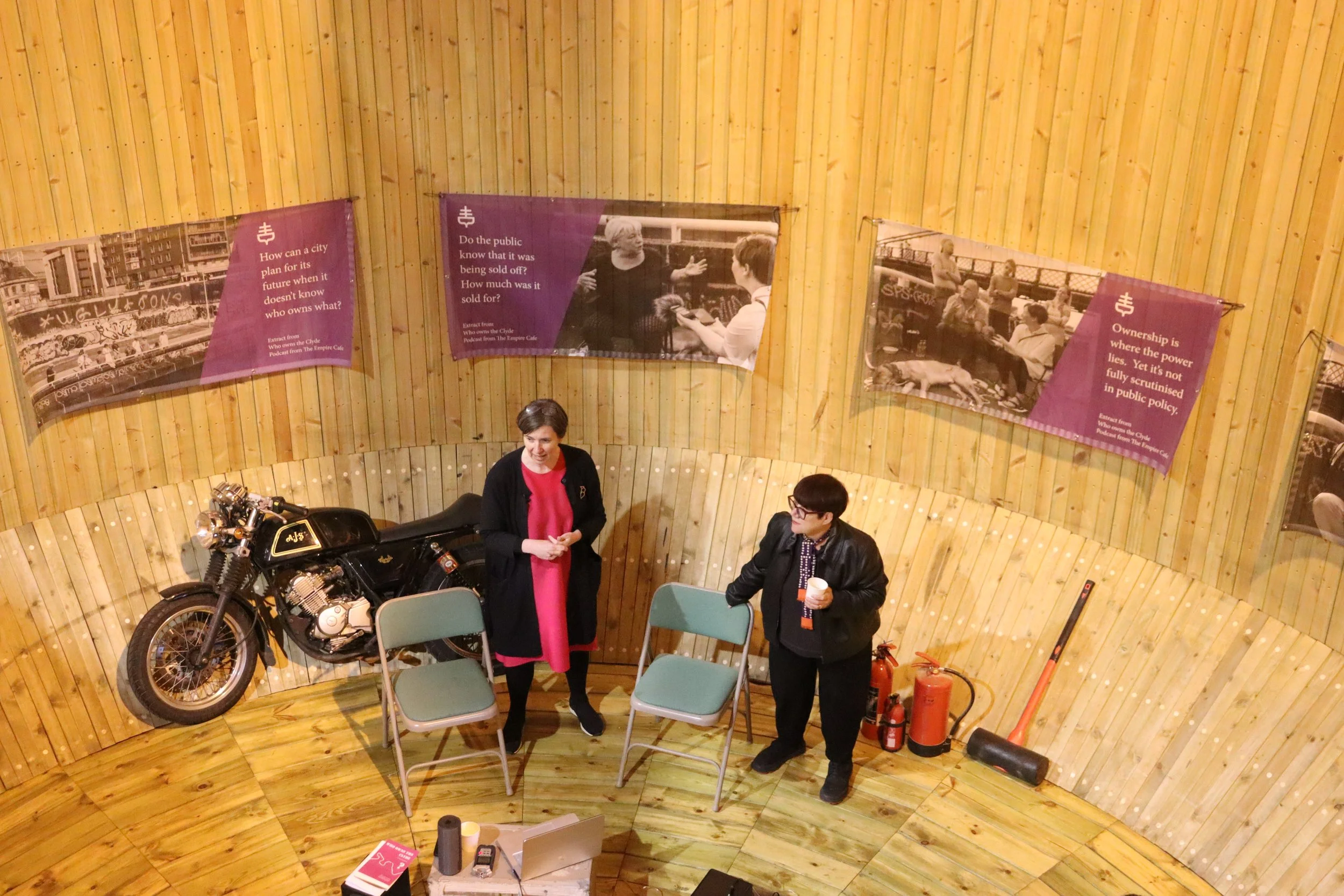 Two people standing and talking in a circular wooden room. The woman on the left is wearing a red dress and a black jacket, and the man on the right is wearing glasses, a black leather jacket, and holding a cup. There are two gray chairs, a vintage black motorcycle mounted on the wall, and some fire extinguishers on the floor. The walls are decorated with large posters with text and images.