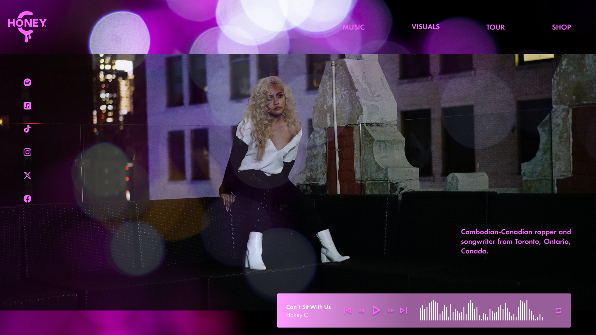 Webpage homepage of Honey, a music artist. Features a woman with curly blonde hair sitting on a black couch outdoors at night, with city buildings in the background. The site includes social media icons on the left and a music player at the bottom di