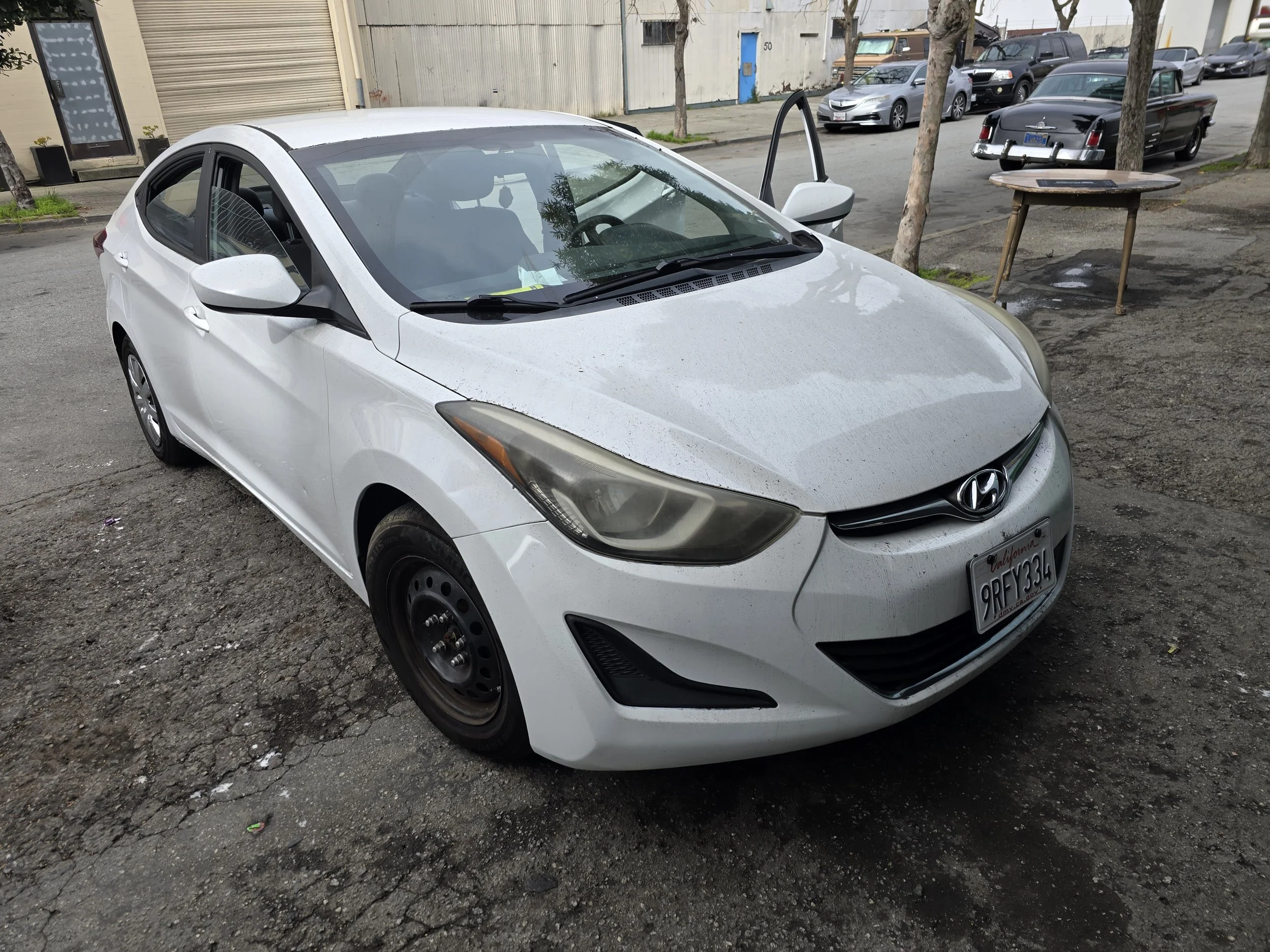 White Hyundai sedan parked on a cracked street, with some dirt and small debris around, and a background of trees, buildings, and other parked cars.