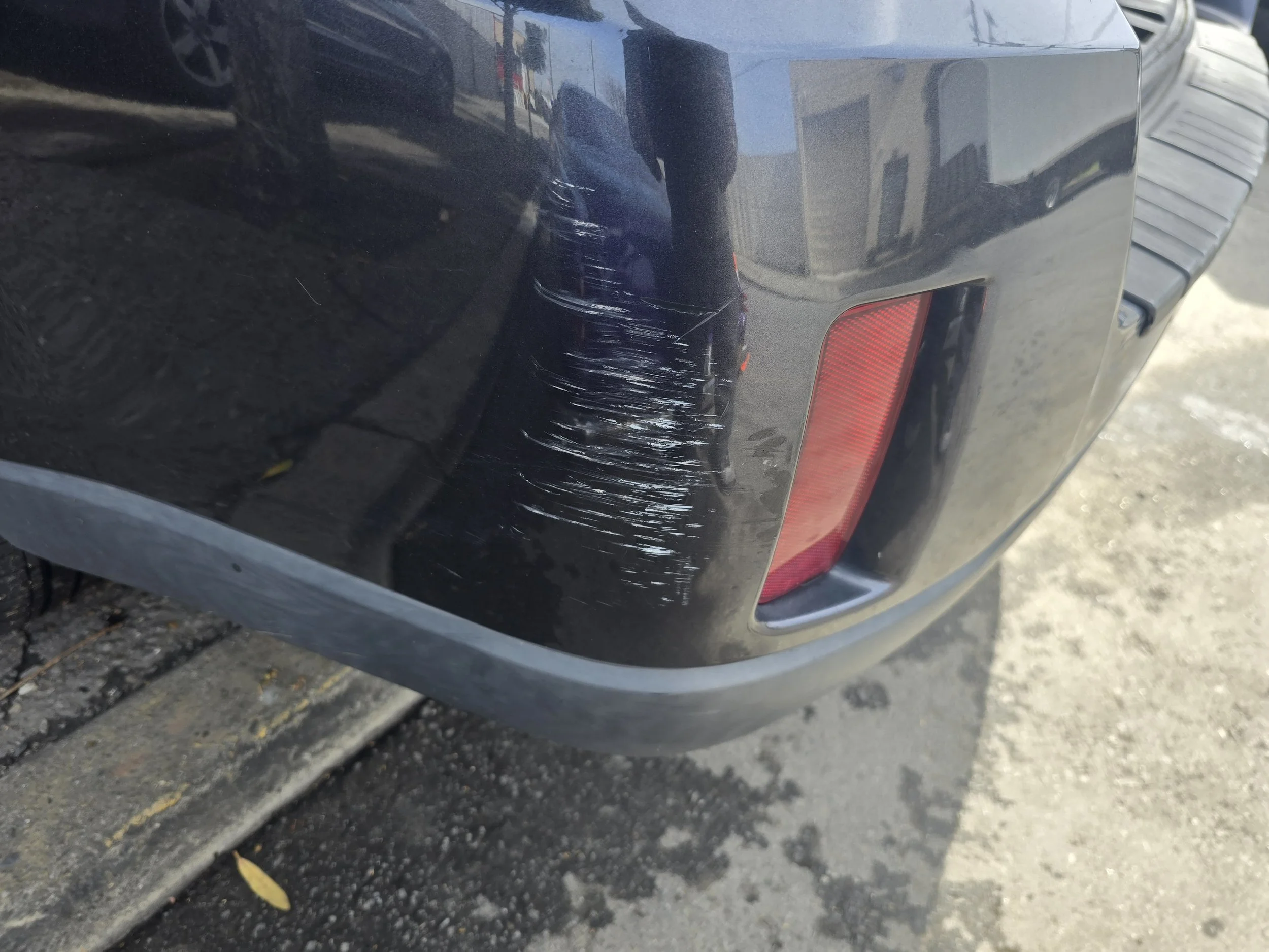 Car bumper with visible scratches and scuffs on the black paint near the red reflector.