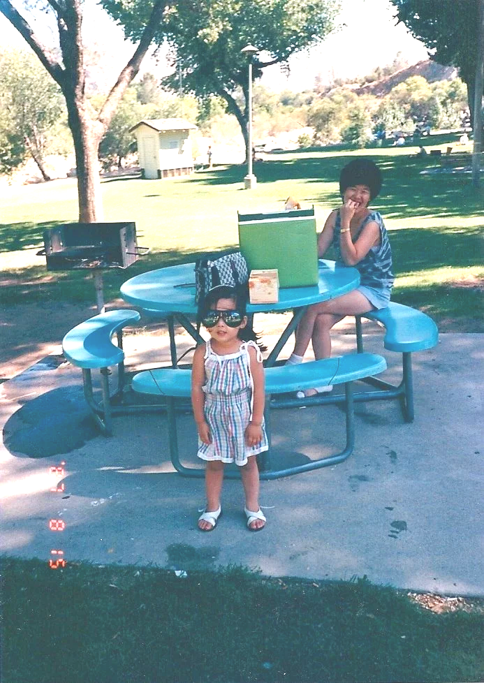 A young girl wearing sunglasses and a striped dress stands in front of a round blue picnic table at a park. A boy sits on the bench of the table, smiling and laughing. There are trees, a small yellow shed, grass, and picnic tables in the background.
