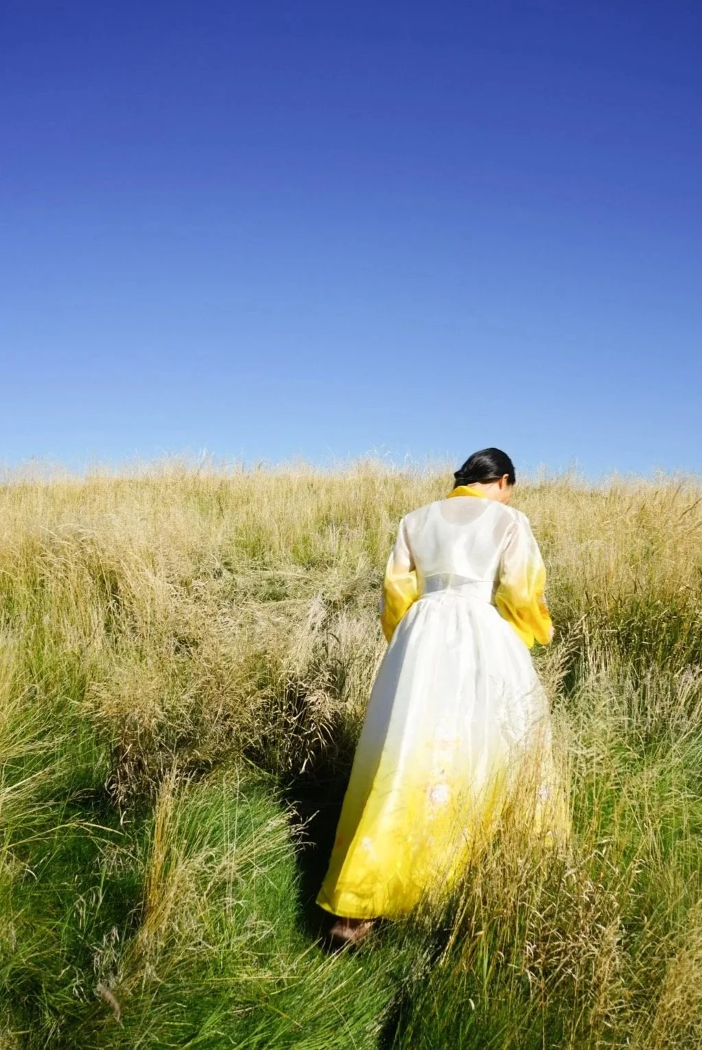 Hannah Abuzaineh, wearing a traditional Korean hanbok, stands with her back turned in a field of tall grass in In Time, a photographic exploration of memory, heritage, and belonging by Here with Han.