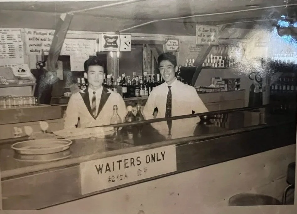 Two young men in white tuxedos behind a bar with bottles and glasses, smiling at the camera. A sign on the bar reads "WAITERS ONLY".