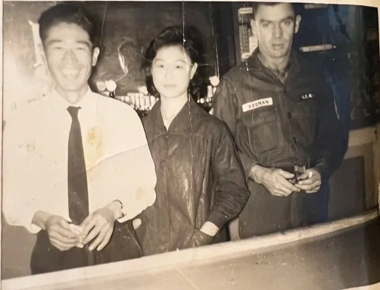 Three people standing at a bar in a vintage black and white photo, smiling or looking serious, with drinks in hand.