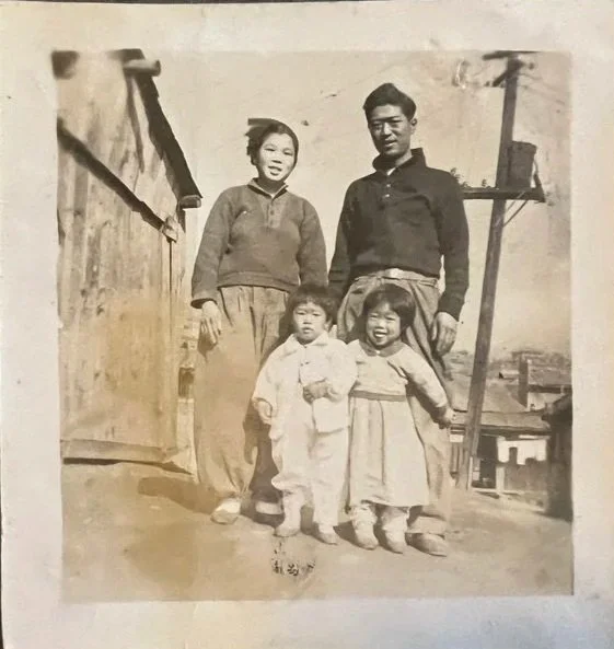 A vintage black and white photo of two adults and two children standing outdoors on a dirt street with wooden structures in the background.