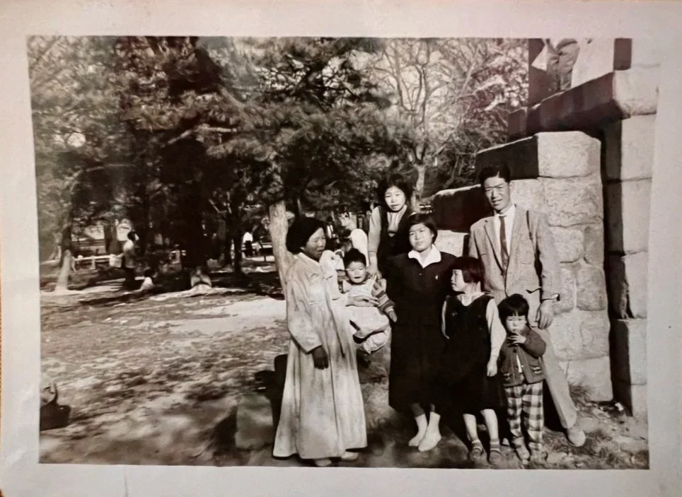 A black-and-white photo of a family of seven standing outdoors near a stone structure and trees. The family includes a man and woman surrounded by four children, some of whom are being held or standing close. The group is dressed in mid-20th century 