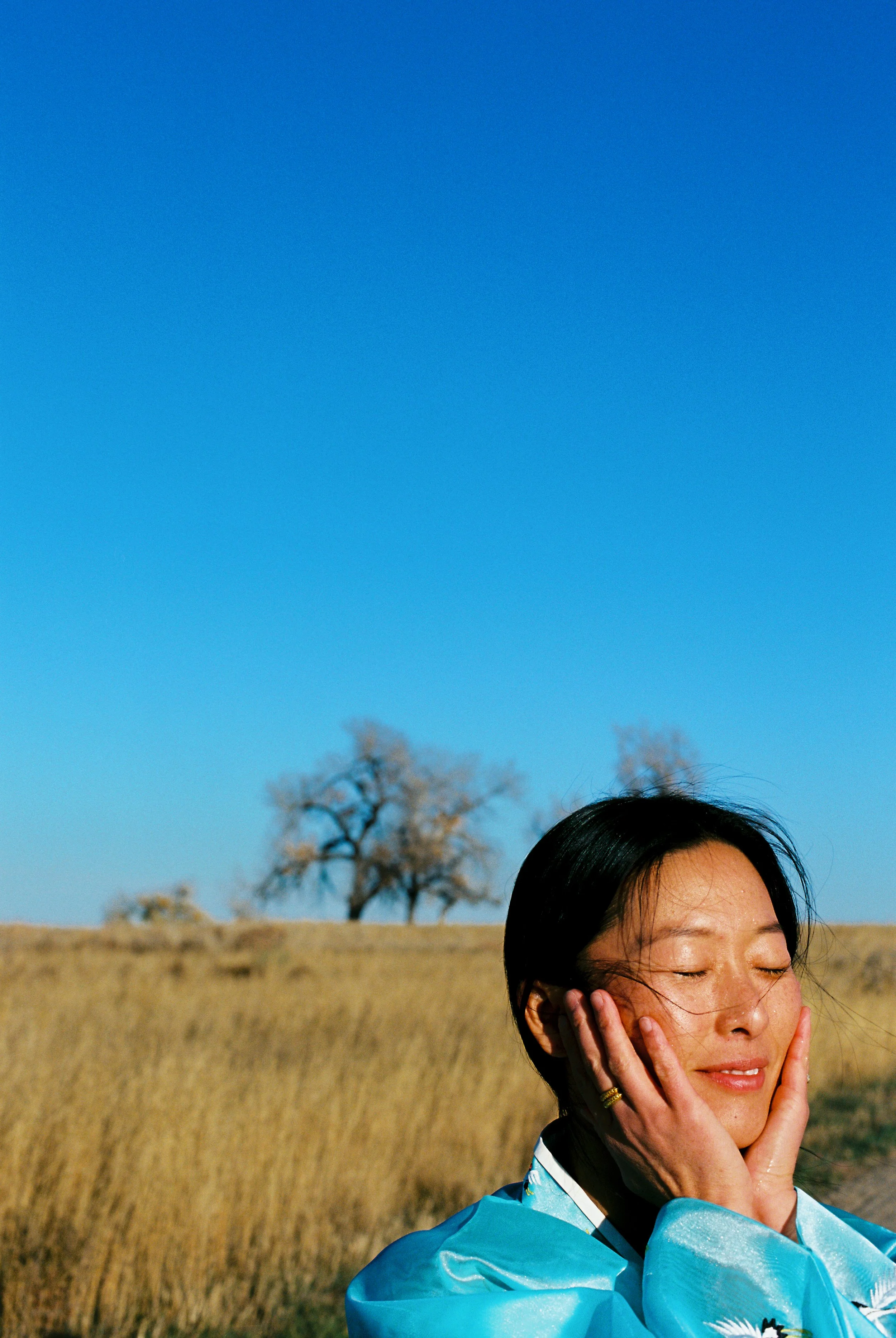 A woman with black hair and fair skin, holding her face with both hands, stands outdoors in a field of dry grass with a tree in the background, under a clear blue sky.