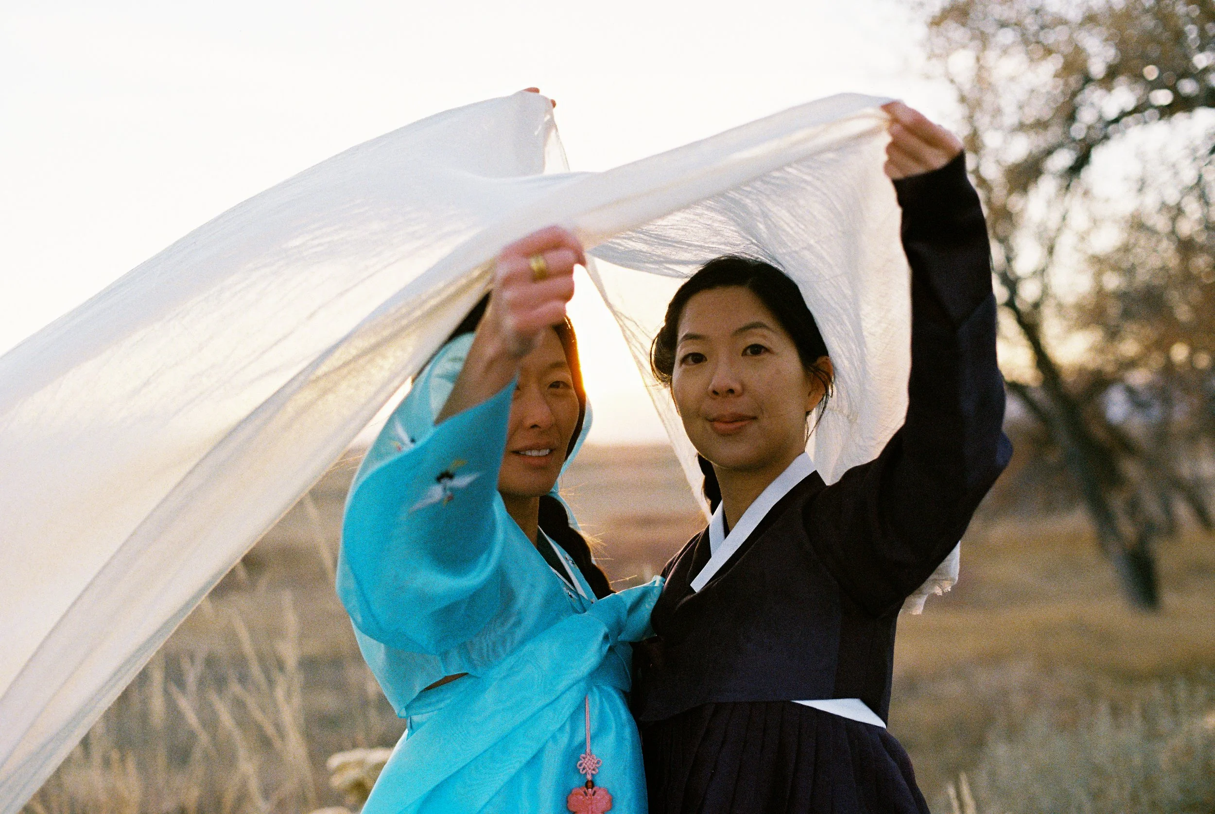 Two women, one in traditional Korean hanbok and the other in modern formal wear, hold a white fabric or paper between them outdoors at sunset or sunrise, with trees in the background.