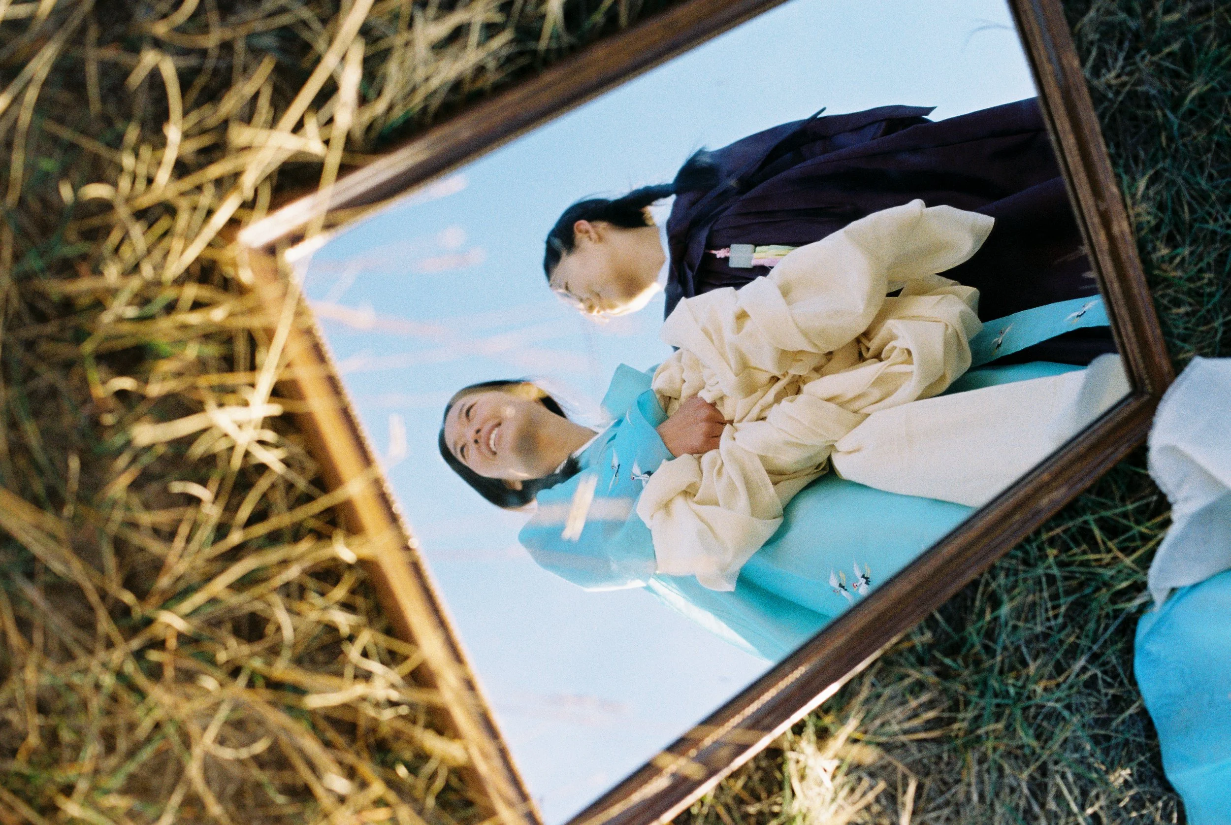 Two children, a boy and a girl, looking at themselves in a mirror placed outdoors in a field of grass and straw, with a clear blue sky in the background.