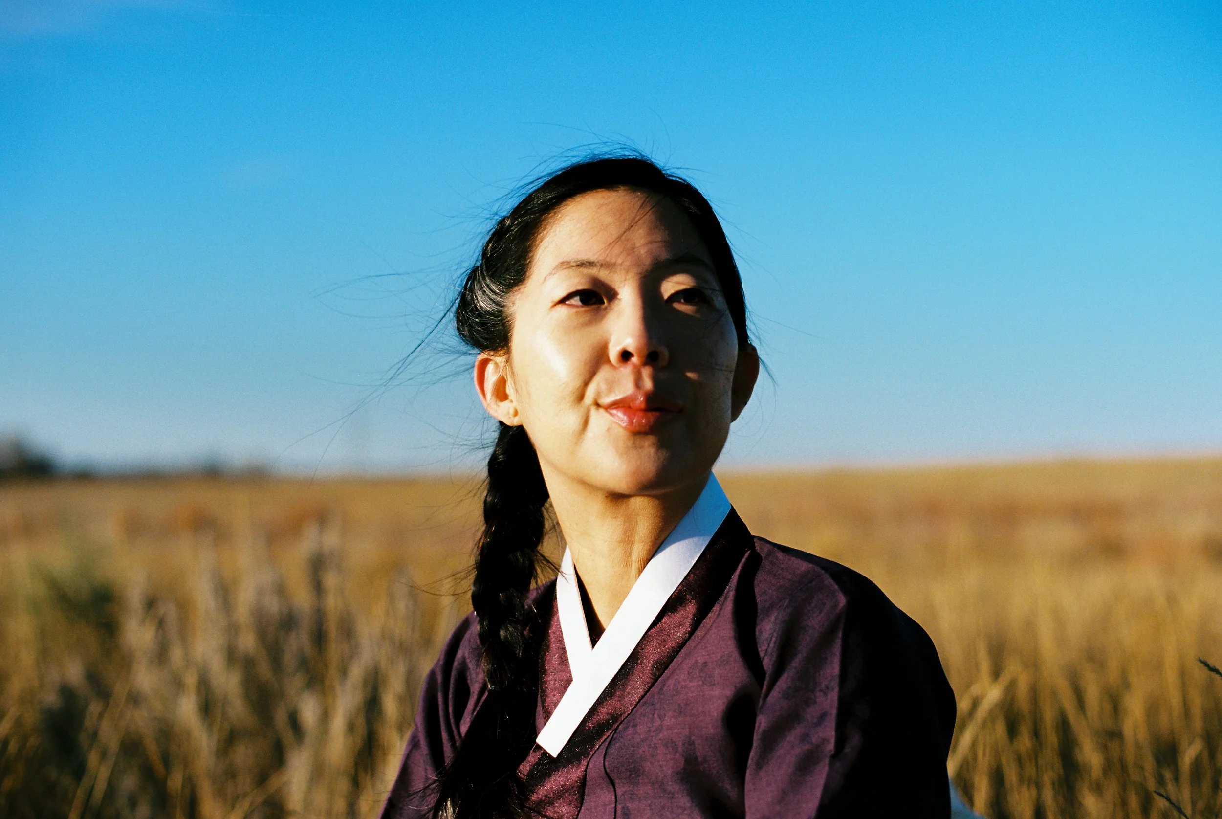 A woman dressed in traditional Korean clothing, standing outdoors in a field with a clear blue sky.