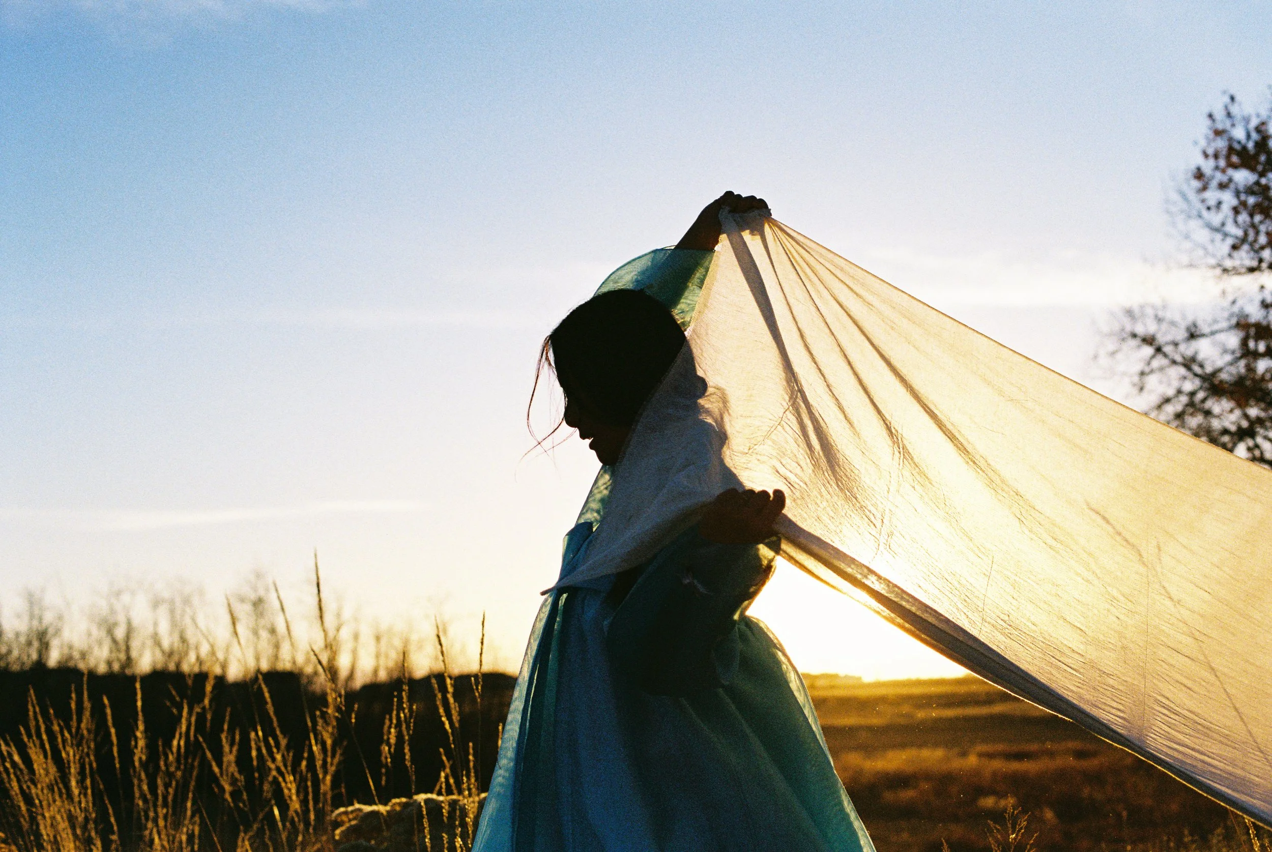 Silhouette of a Hannah Abuzaineh carrying a yellow tent against the backdrop of a sunset or sunrise in a rural area with tall grass and trees.
