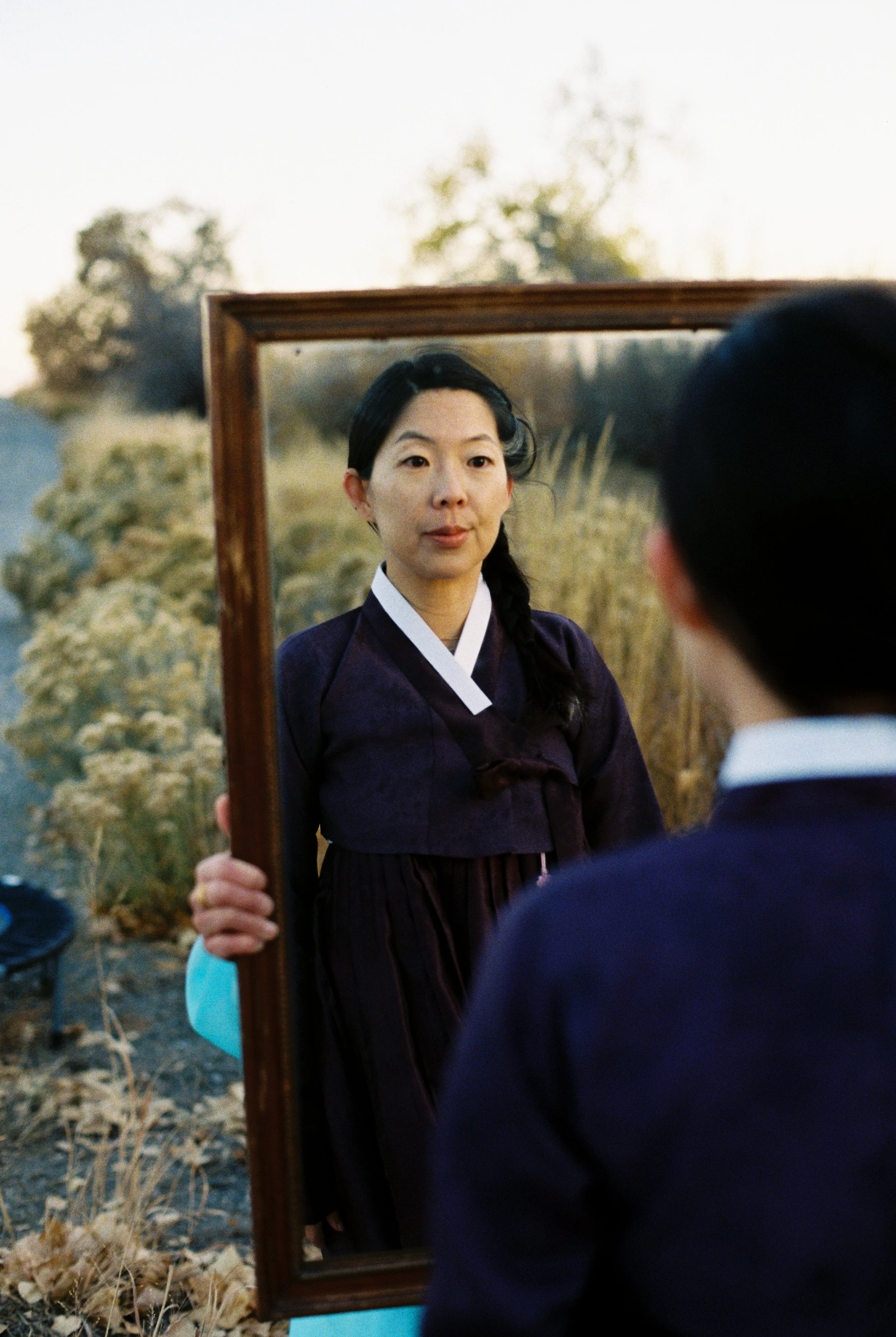 A woman dressed in traditional Korean attire looking at her reflection in a mirror, outdoors with trees and dried grass in the background.