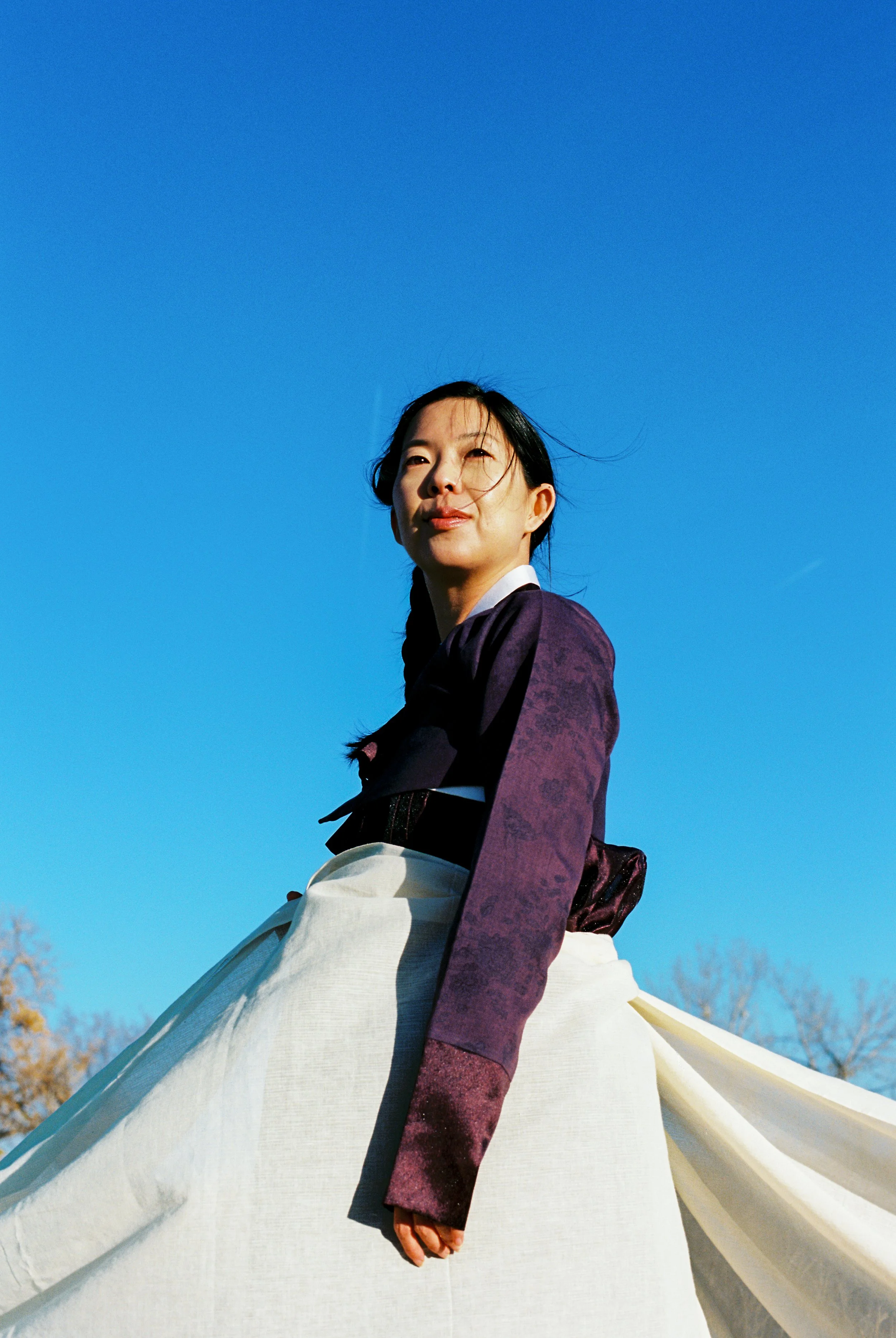 A woman with black hair pulled back, wearing a dark purple kimono with floral patterns, standing outdoors under a clear blue sky, with trees in the background.