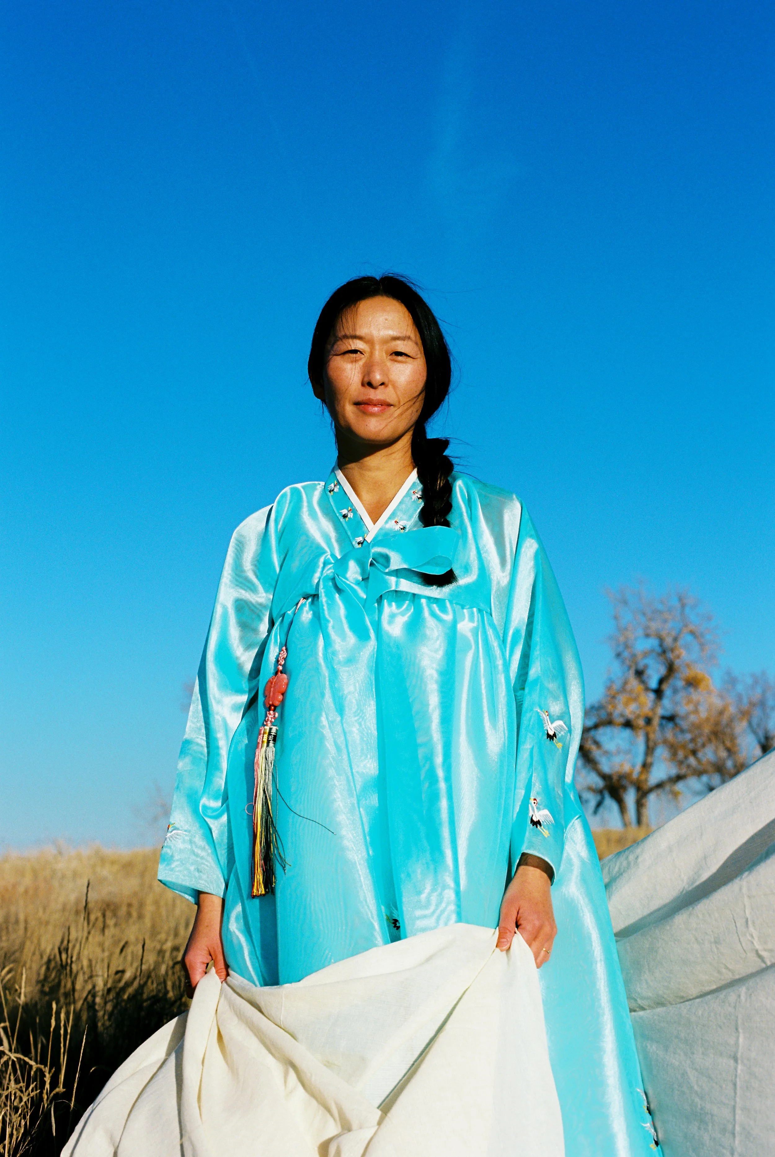 A woman in a shiny light blue traditional Asian dress with embroidered cranes and a bow, standing outdoors in a field with tall grasses, under a clear blue sky with a leafless tree in the background.