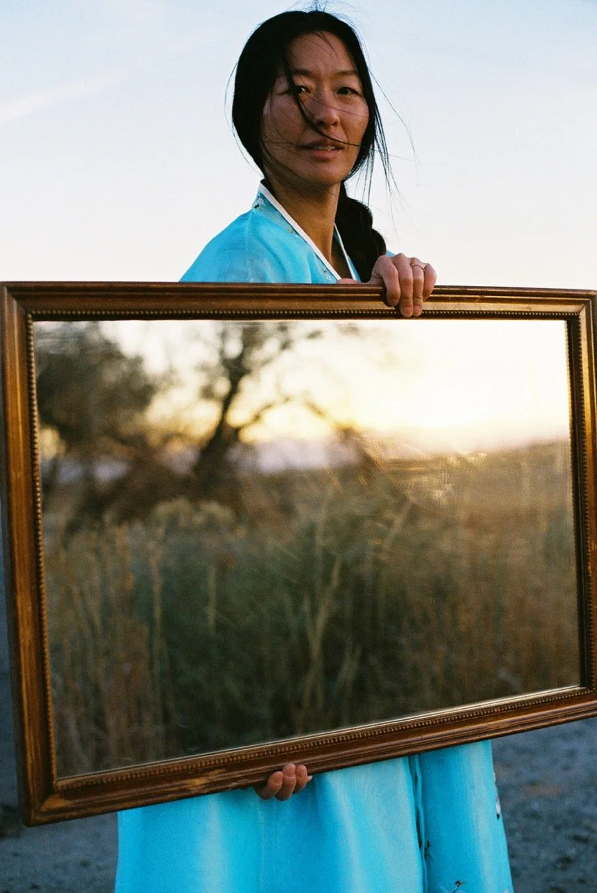 A woman with long black hair wearing a blue top holds a large framed mirror outdoors during sunset, with trees and sky reflected in the mirror.