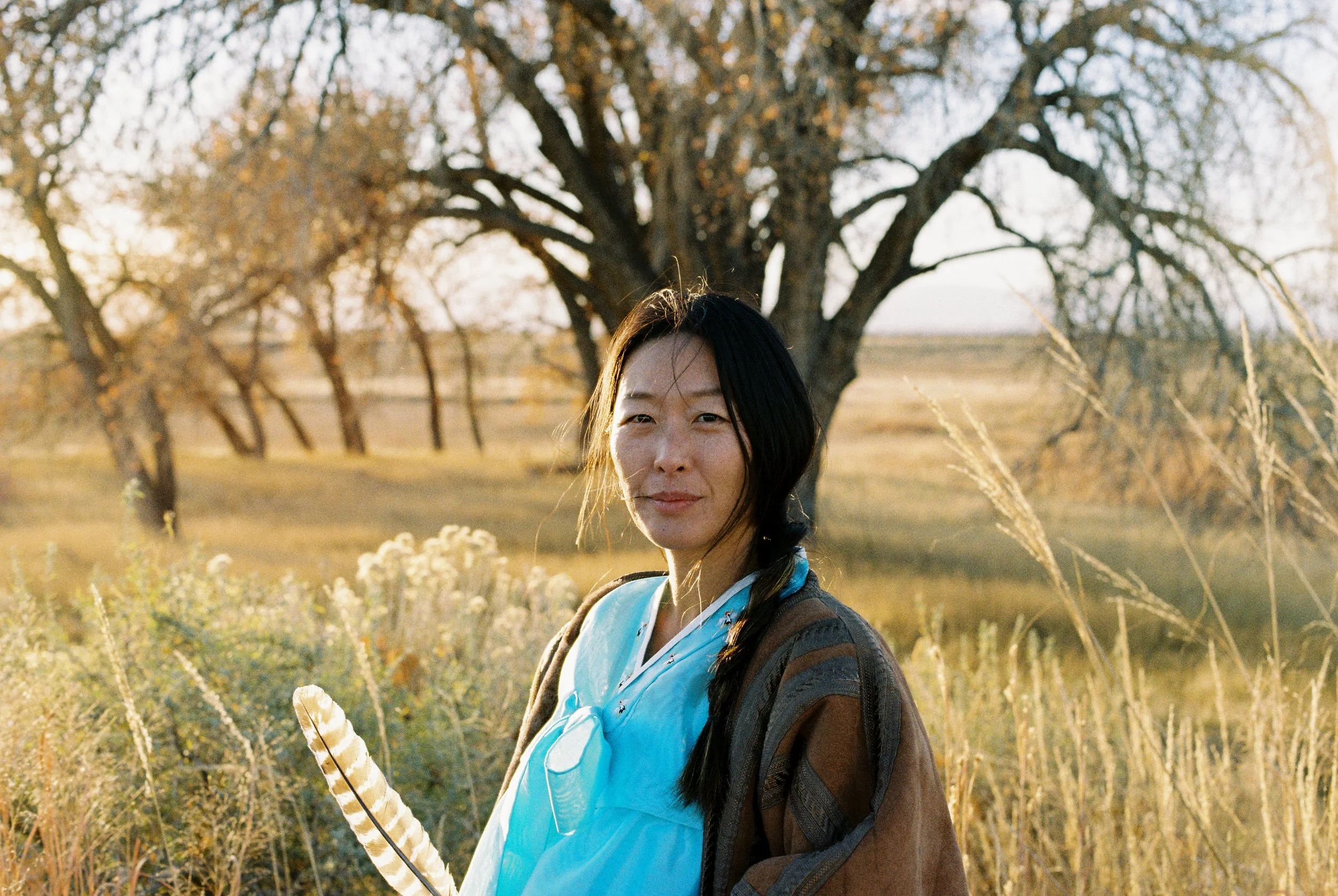 A woman dressed in traditional Japanese attire standing outdoors with autumn trees in the background.
