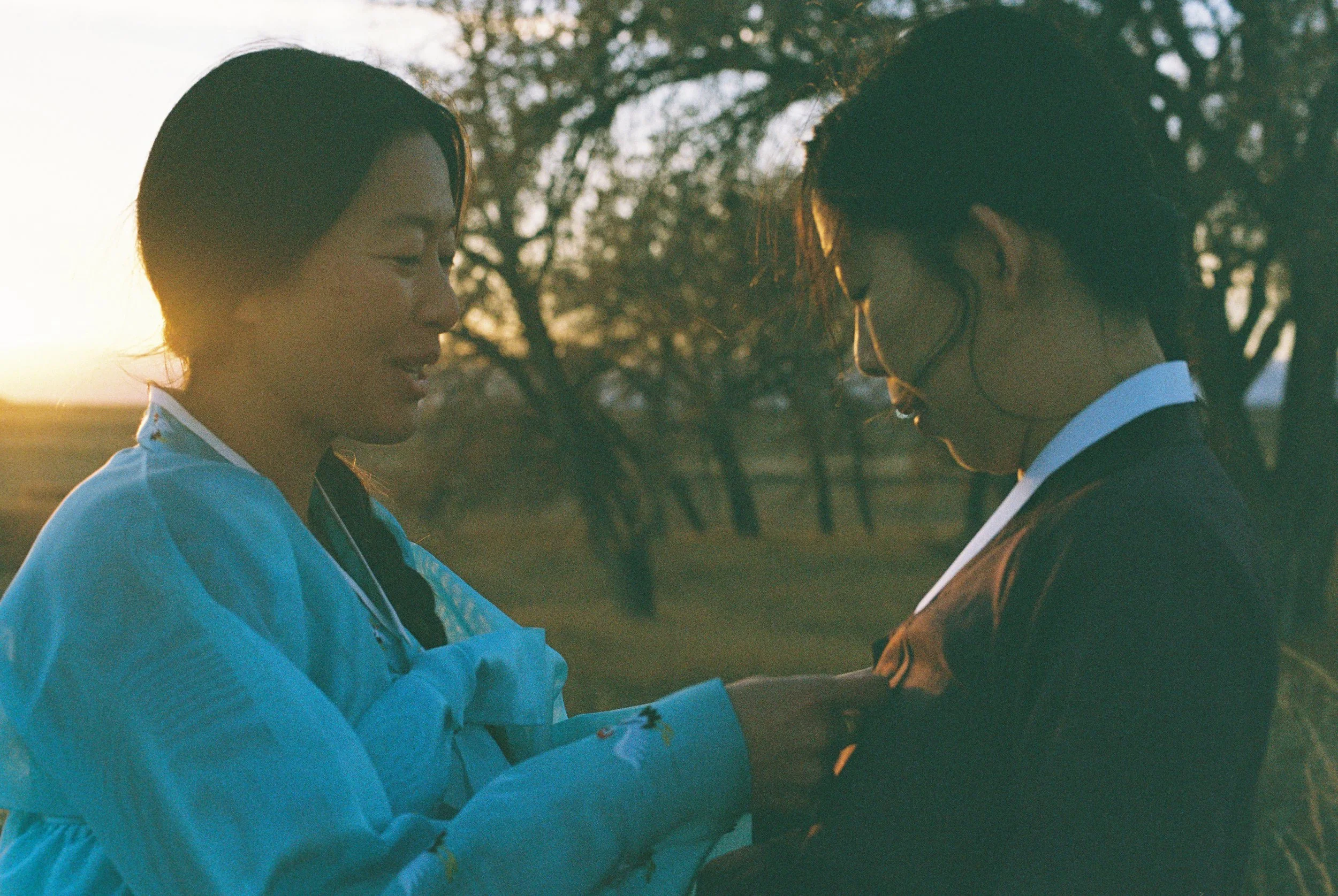 Two women in light blue clothing standing outdoors at sunset, facing each other with their eyes closed and hands touching, in a tender moment.