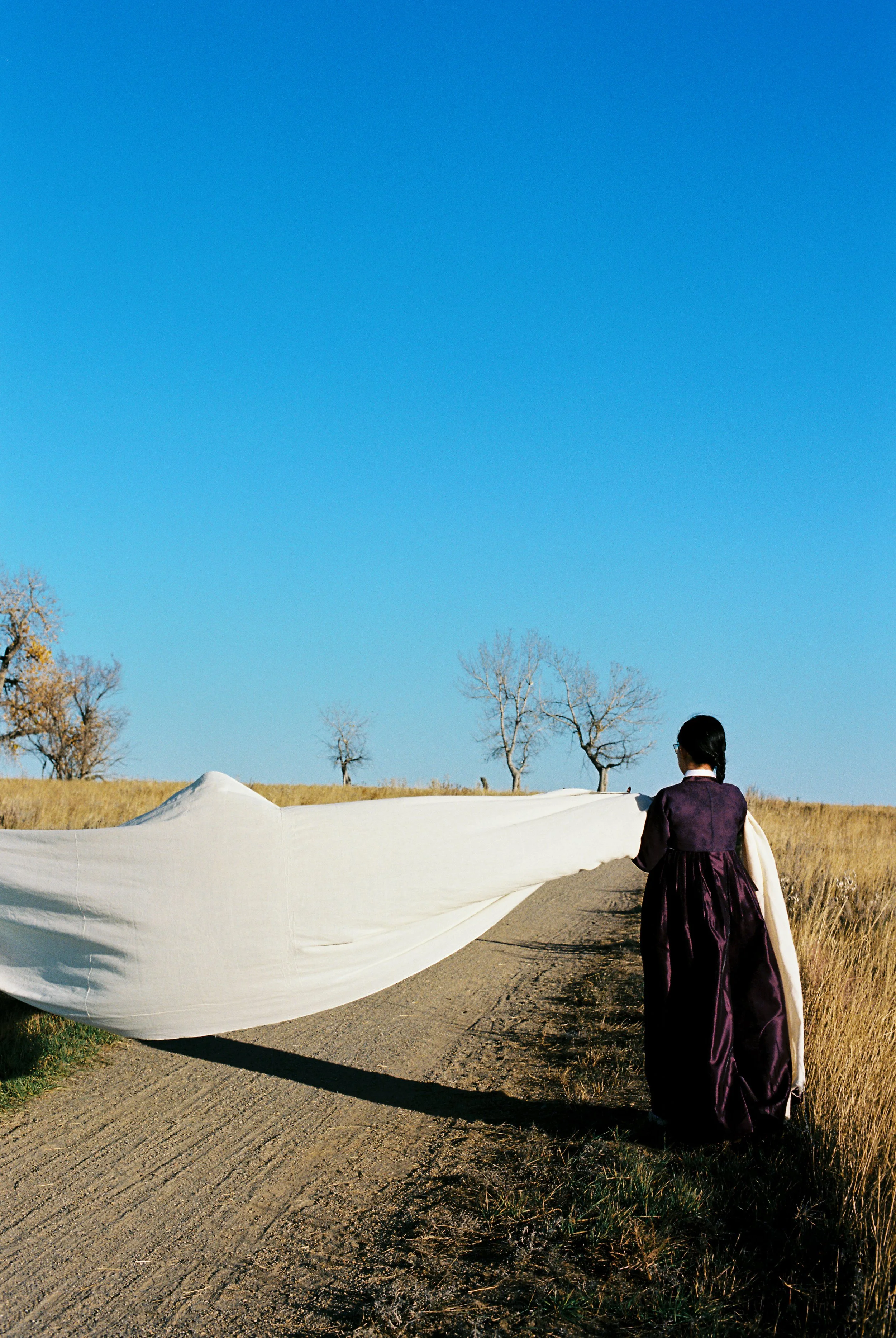 Grace Kim dressed in traditional clothing walking on a dirt path in a rural area with grass and leafless trees, holding a large white fabric or sheet.