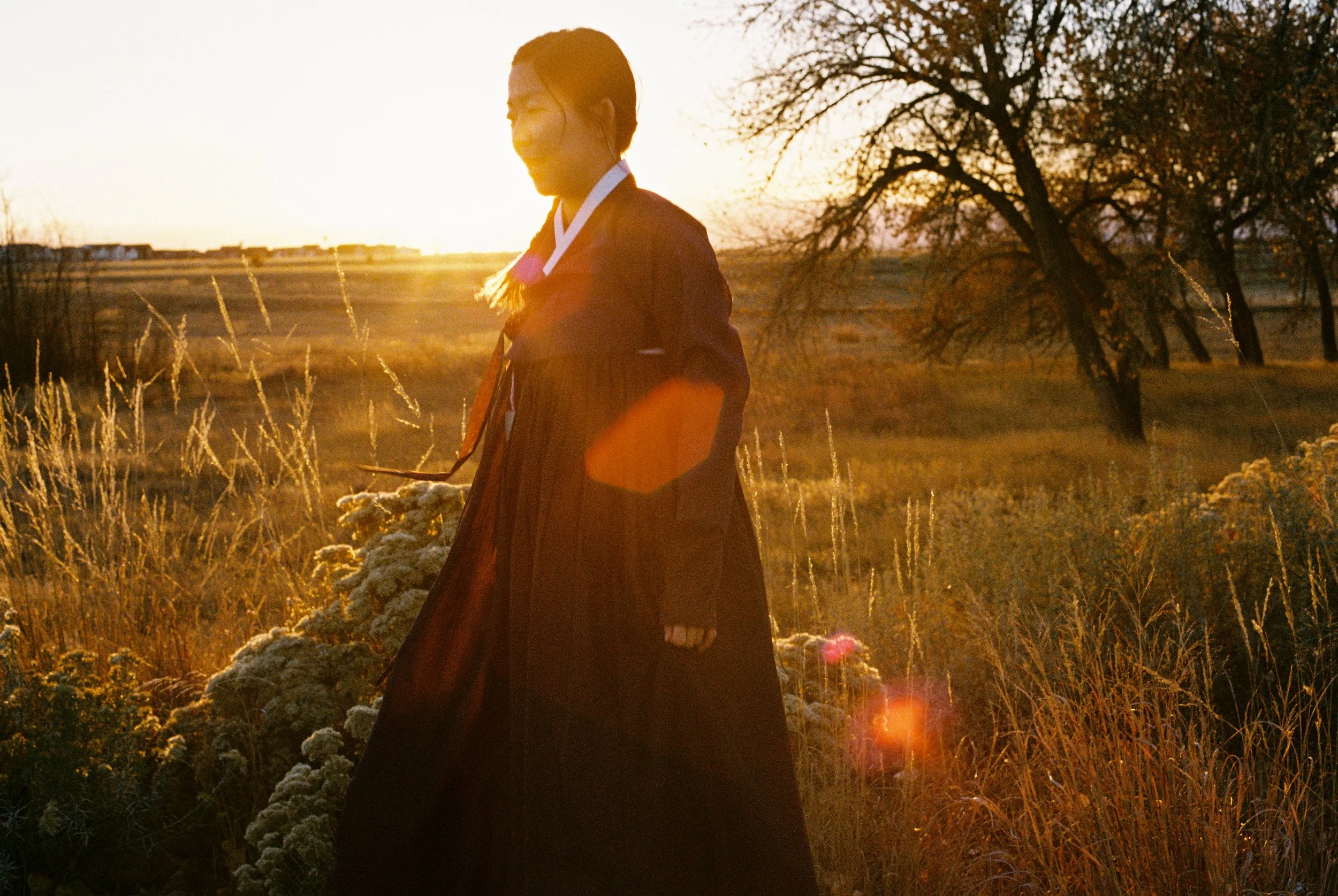 A woman in a long dress walking outdoors during sunset, with a tree and open fields in the background.