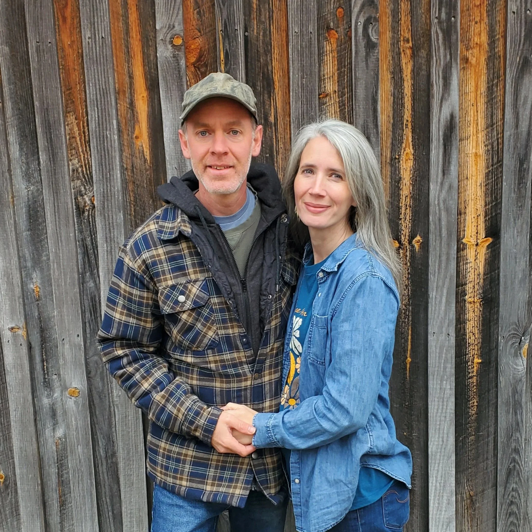 A man and woman standing close together, holding hands and smiling in front of a wooden fence.