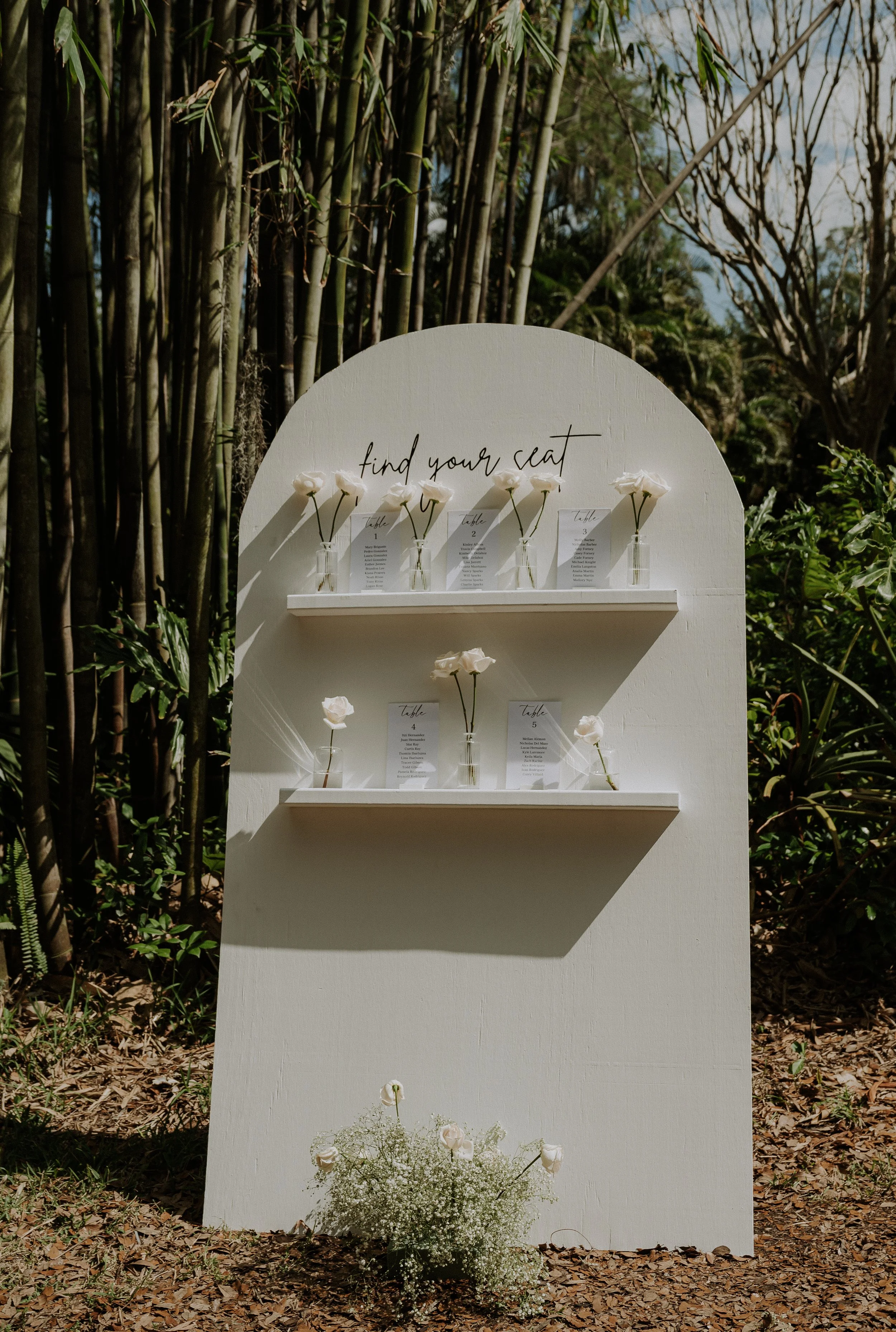 A white wedding seat chart with small bottles of white roses on two shelves, set outdoors among trees and greenery.