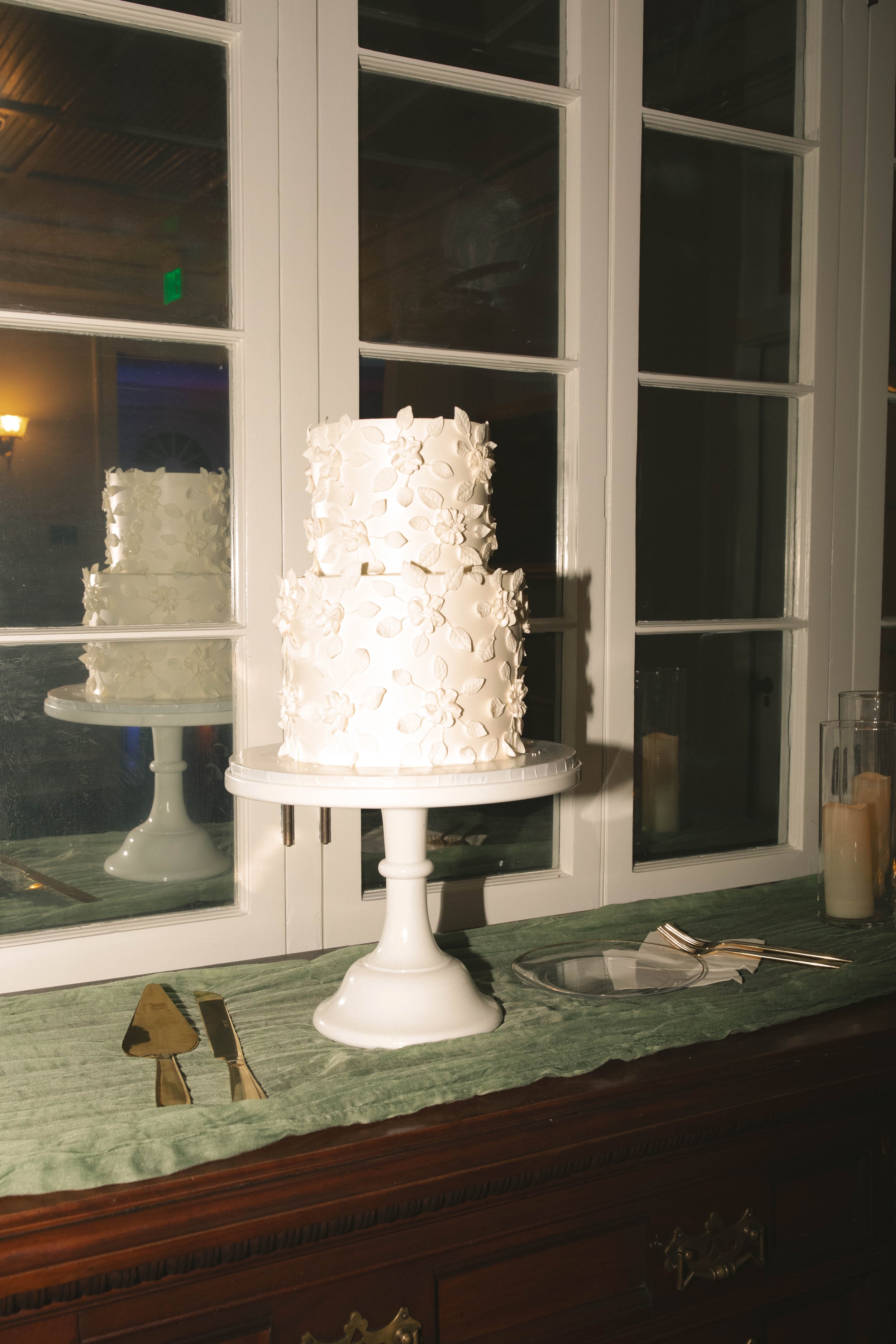 A two-tiered white wedding cake with floral decorations on a white cake stand. The cake is placed on a table with a green cloth, with a cake knife, server, a plate, and two forks nearby, in front of a window with reflections of the cake.