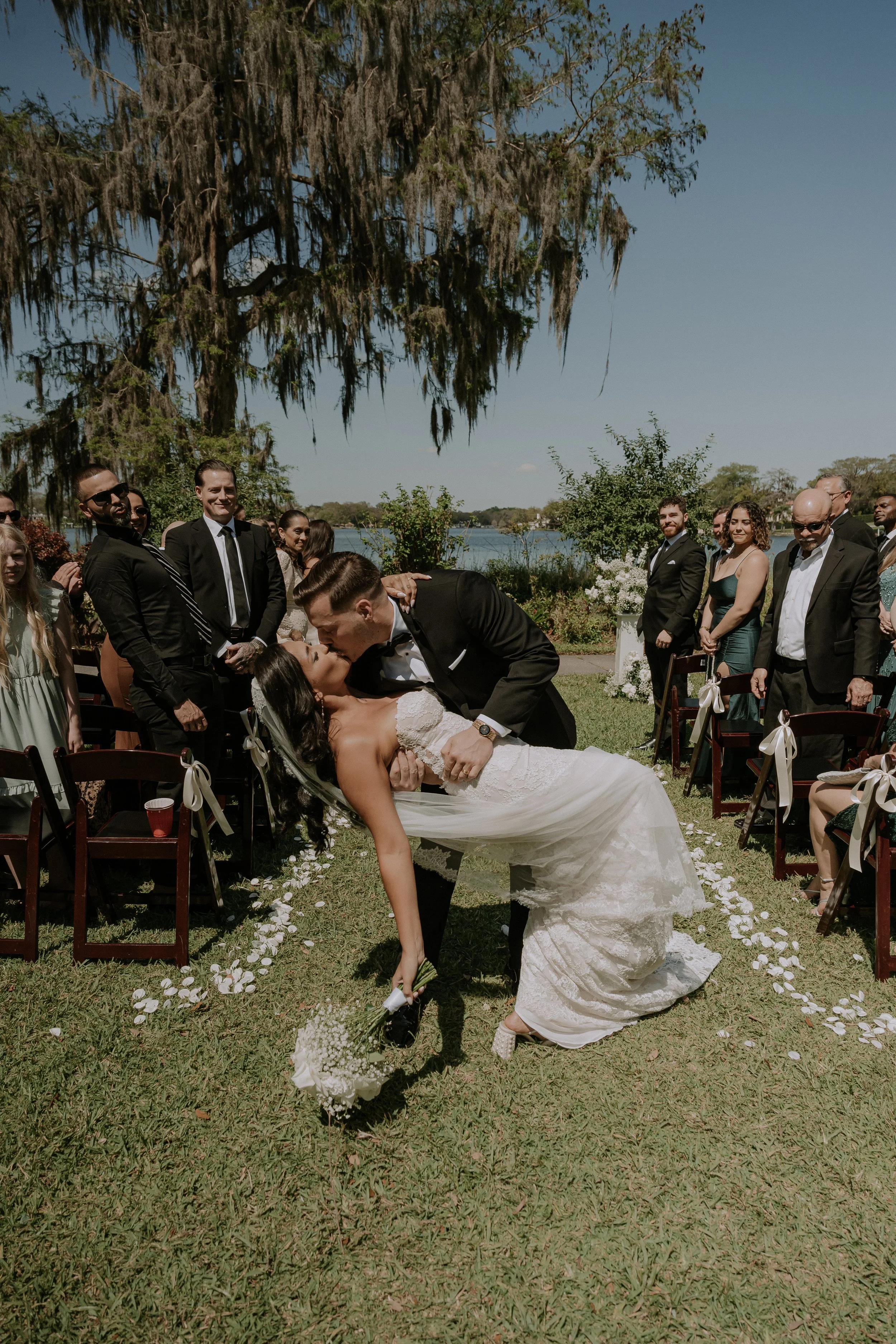 A newlywed couple sharing a romantic kiss outdoors during their wedding ceremony, with guests seated on either side under a large tree, by a lakeside on a sunny day.