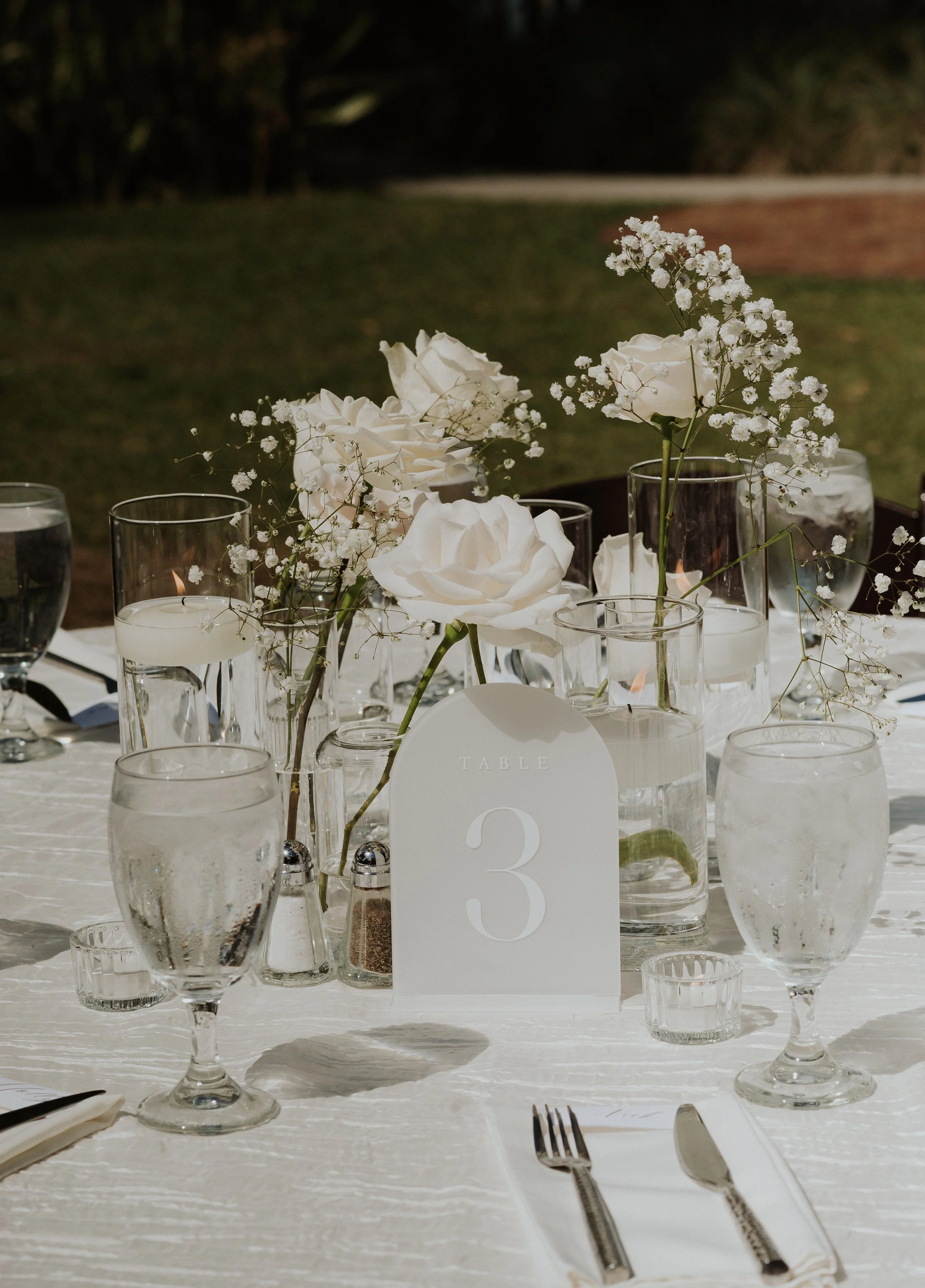 Elegant outdoor table setting with white floral centerpiece, candles, and glassware, with a table number card indicating table 3.