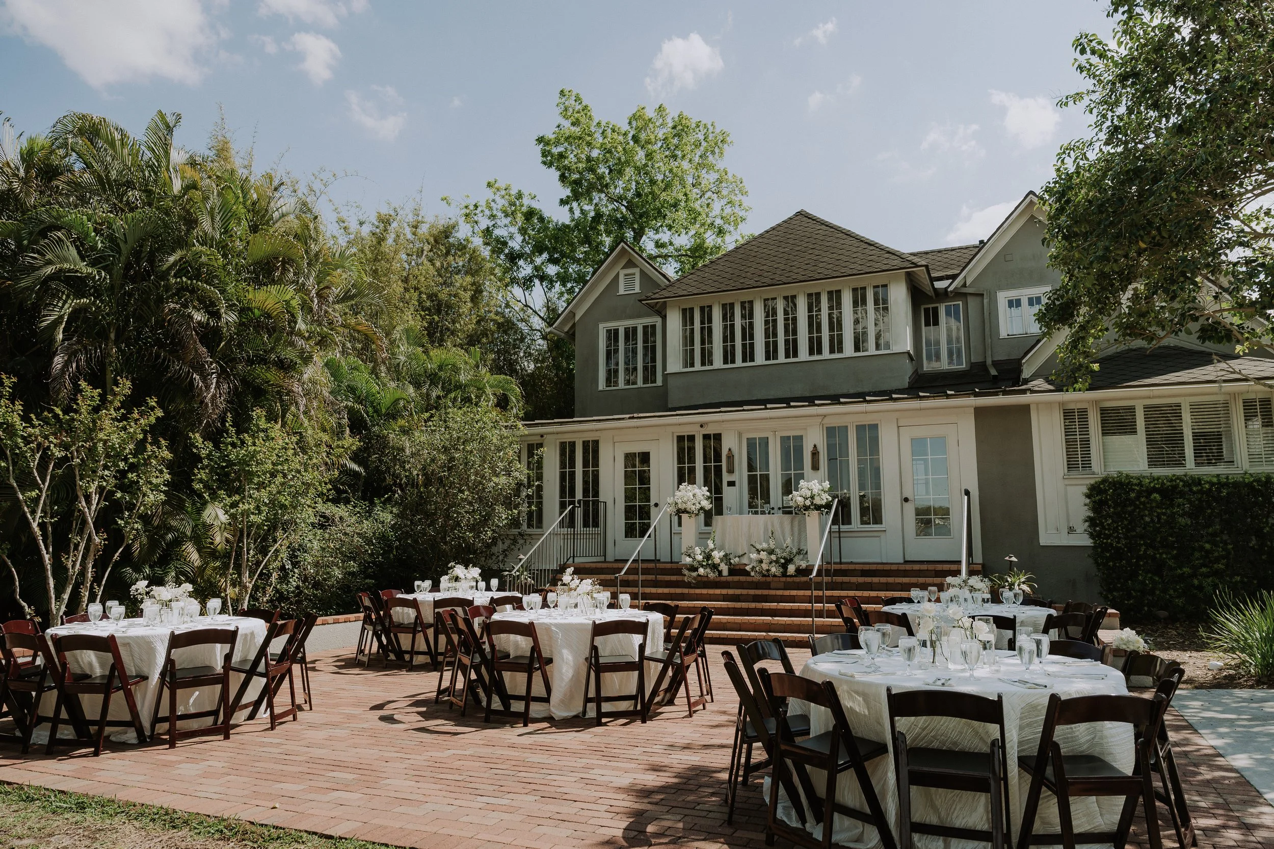 Outdoor wedding or event setup in a backyard or garden area with tables, chairs, and decorated with white flowers, in front of a large gray house with multiple windows and steps leading up to the door, surrounded by lush green trees and shrubs.