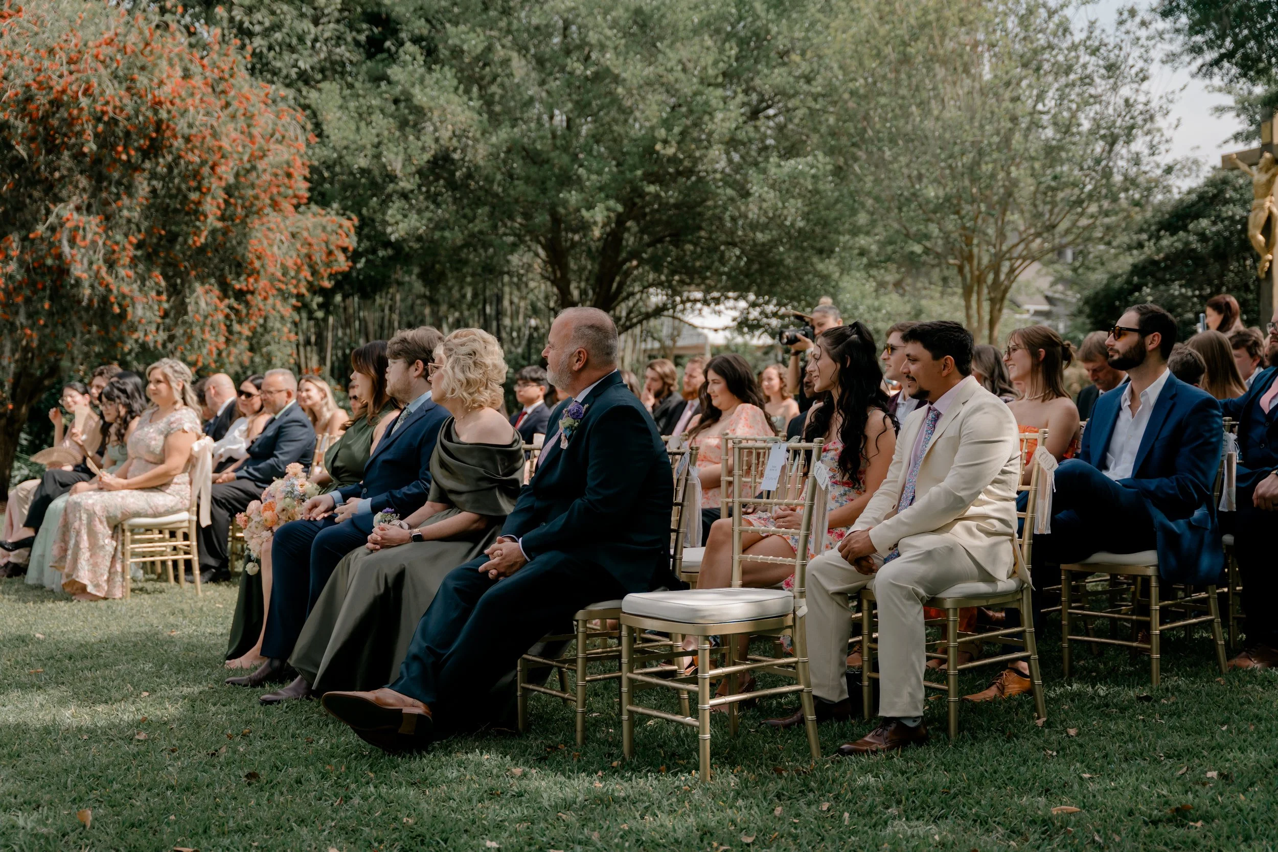 People sitting on chairs outdoors, attending a wedding or formal event, with trees and greenery in the background.