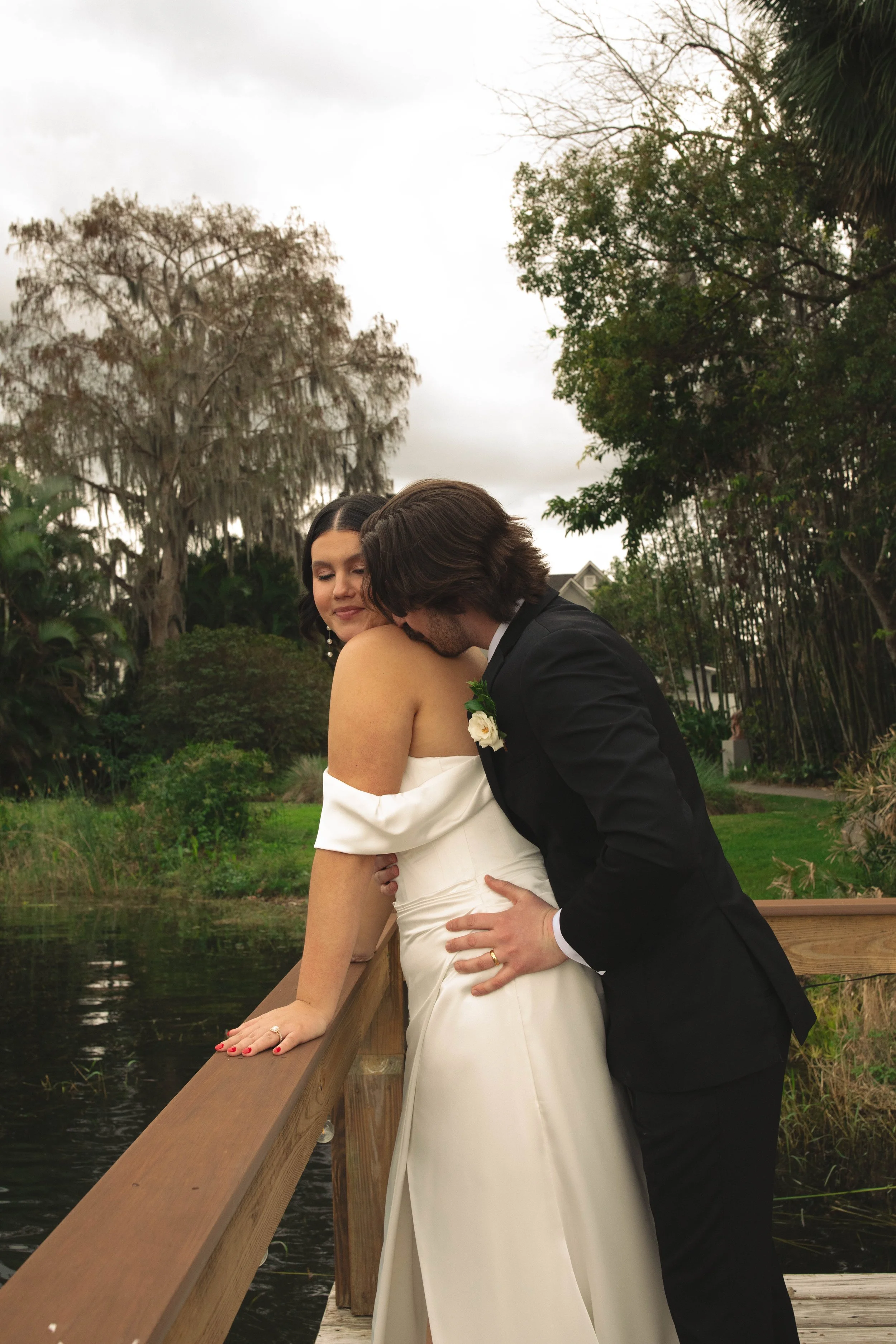 A bride and groom sharing a kiss on a wooden bridge over a pond, surrounded by trees and greenery during a cloudy day.