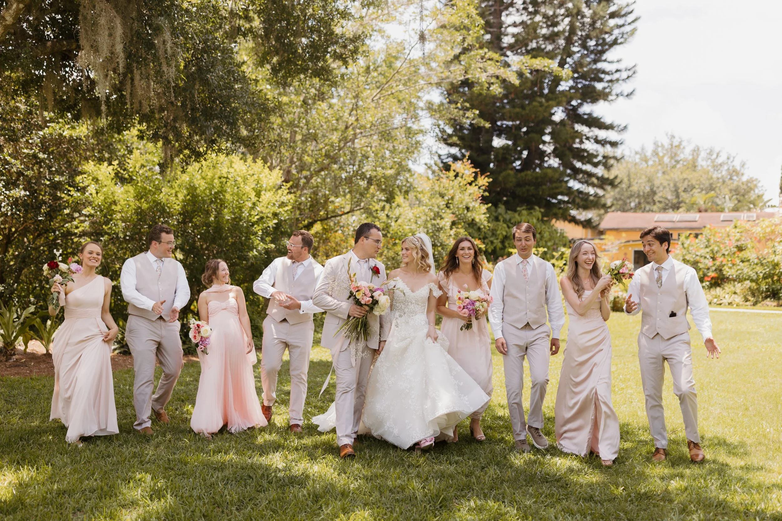 Always Events Co  Orlando weddiA wedding party walking on grass in a park-like setting with trees, consisting of a bride in a white gown, bridesmaids in pastel pink dresses holding bouquets, and groomsmen in light-colored suits, enjoying a sunny day.