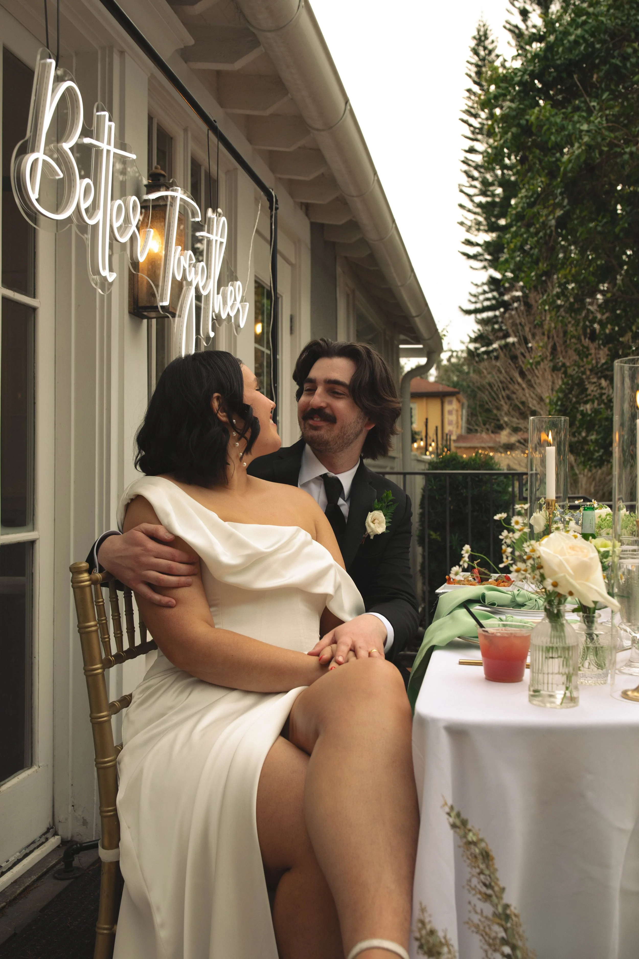 A newlywed couple sharing a moment at their wedding reception, seated at a decorated table on a patio with a sign reading 'Better Together' in the background.