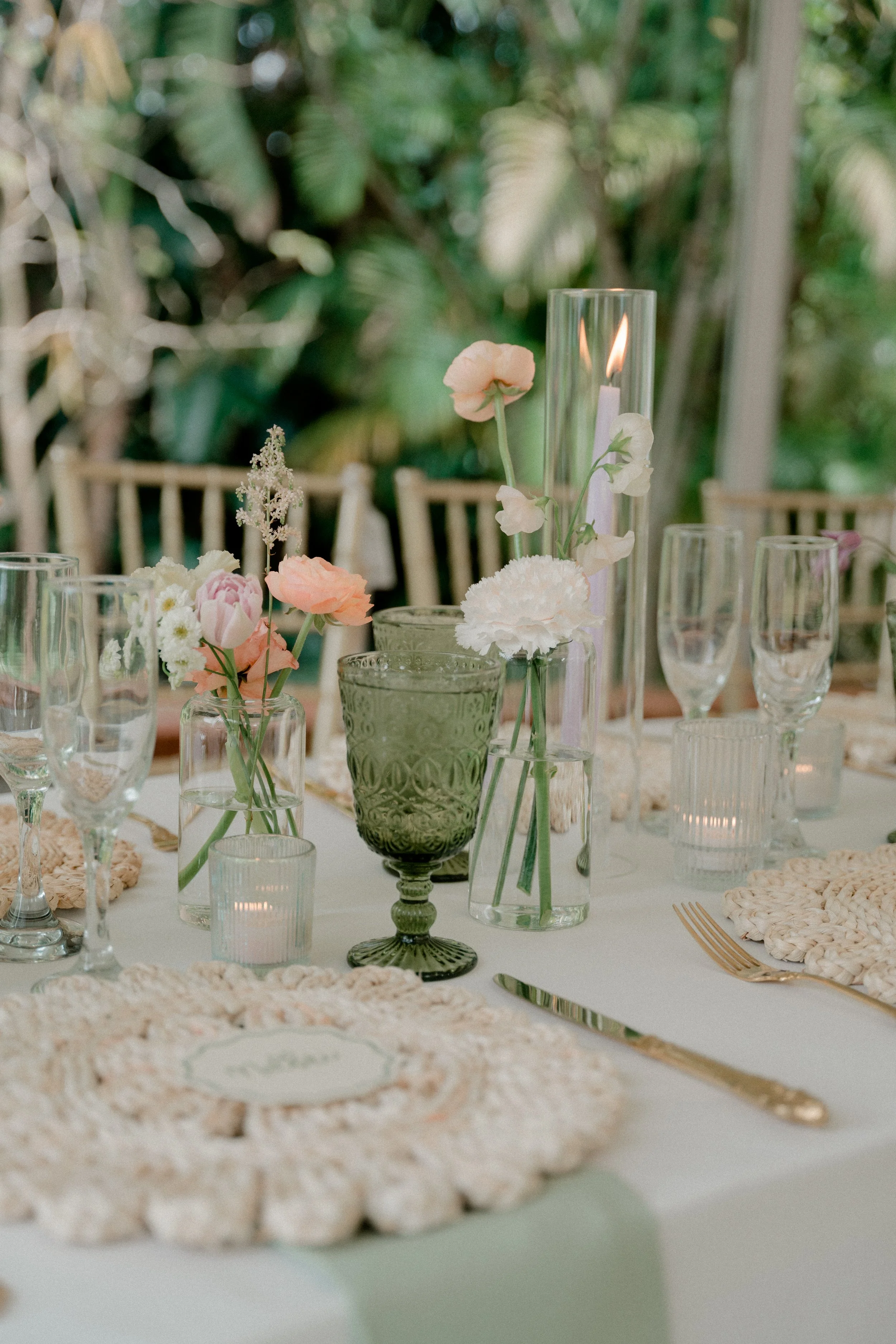 Elegant table setting with pastel pink and white flowers in glass vases, surrounded by candles in glasses, with gold cutlery, set on a white tablecloth in a lush green background.