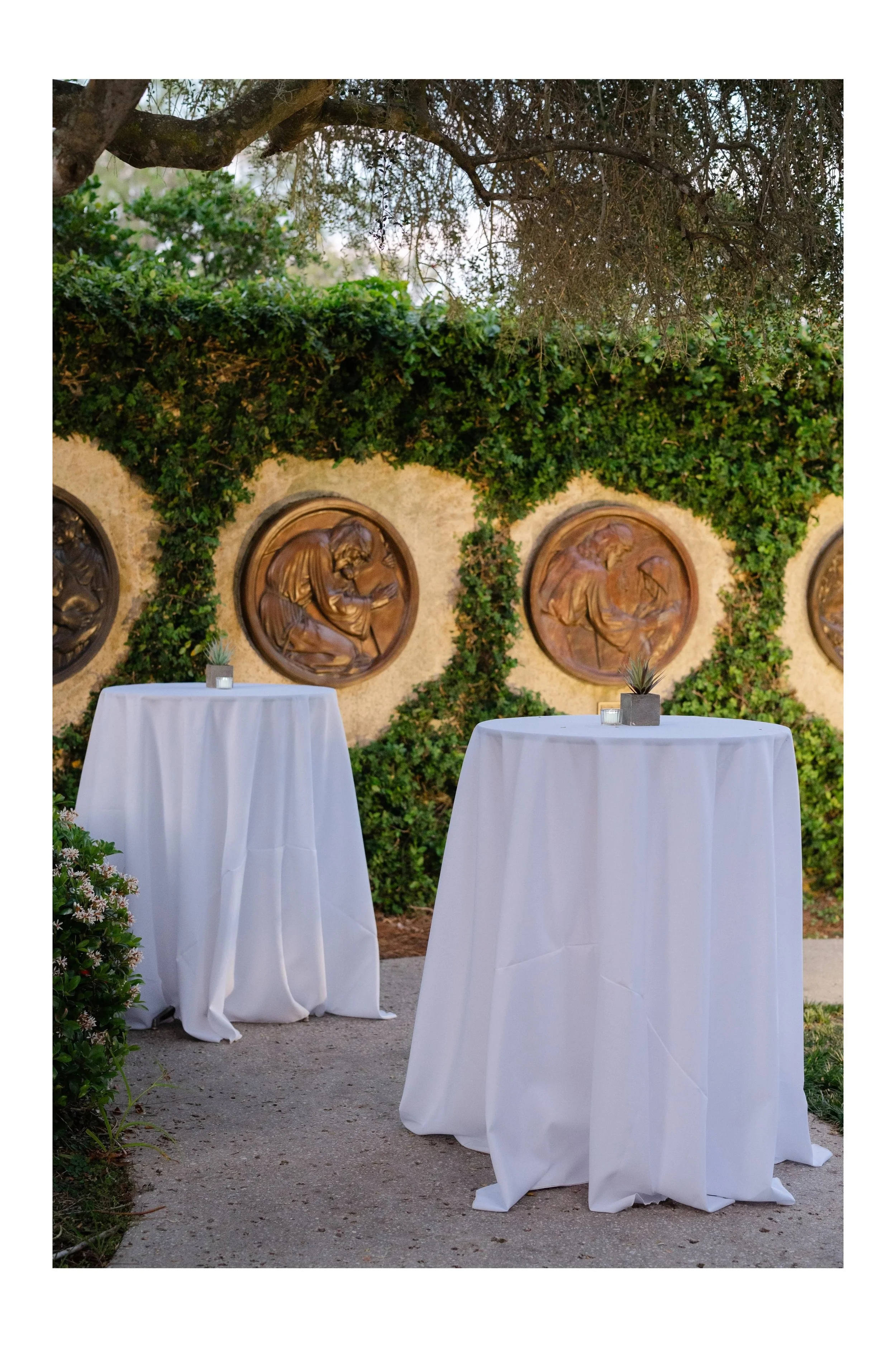 Two round cocktail tables with white tablecloths, each with a small potted plant and candle, set outdoors against a stone wall decorated with bronze relief sculptures and surrounded by green foliage and trees.
