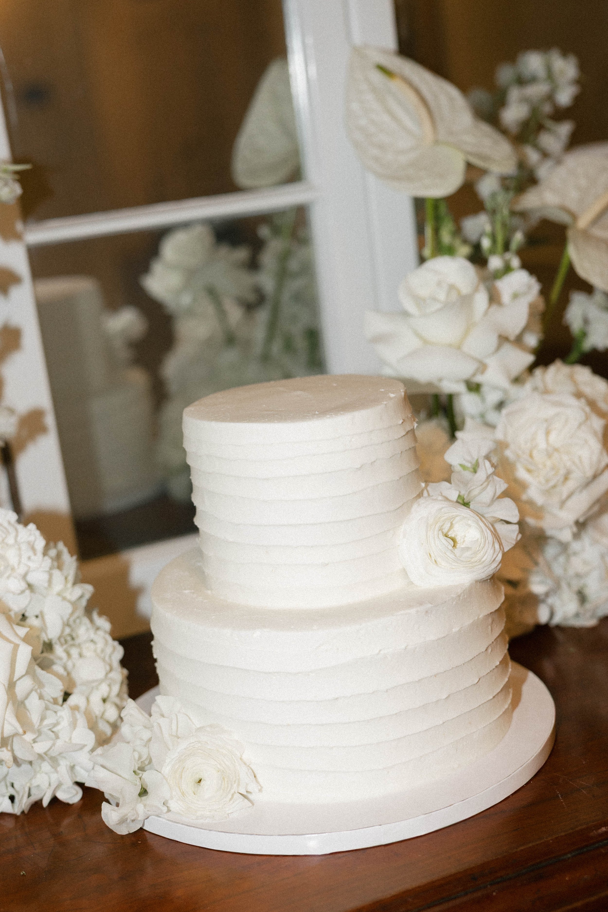 Two-tiered white wedding cake decorated with white flowers on a wooden surface surrounded by white floral arrangements.