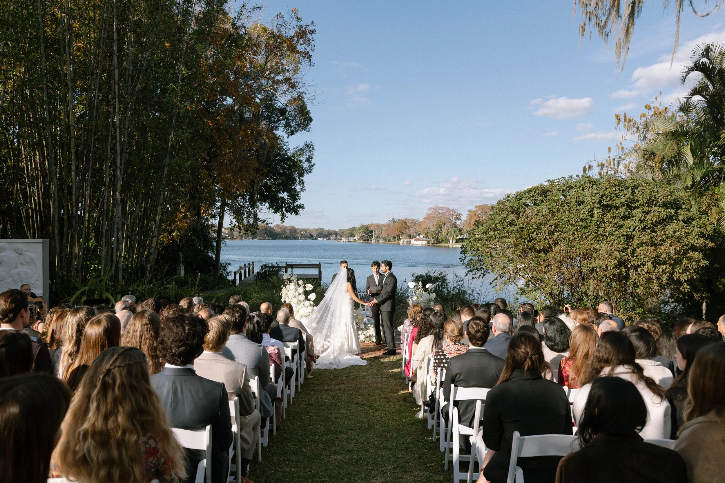 A wedding ceremony outdoors near a lake with guests seated on white chairs, the bride and groom holding hands and exchanging vows, surrounded by trees and flowers on a sunny day.