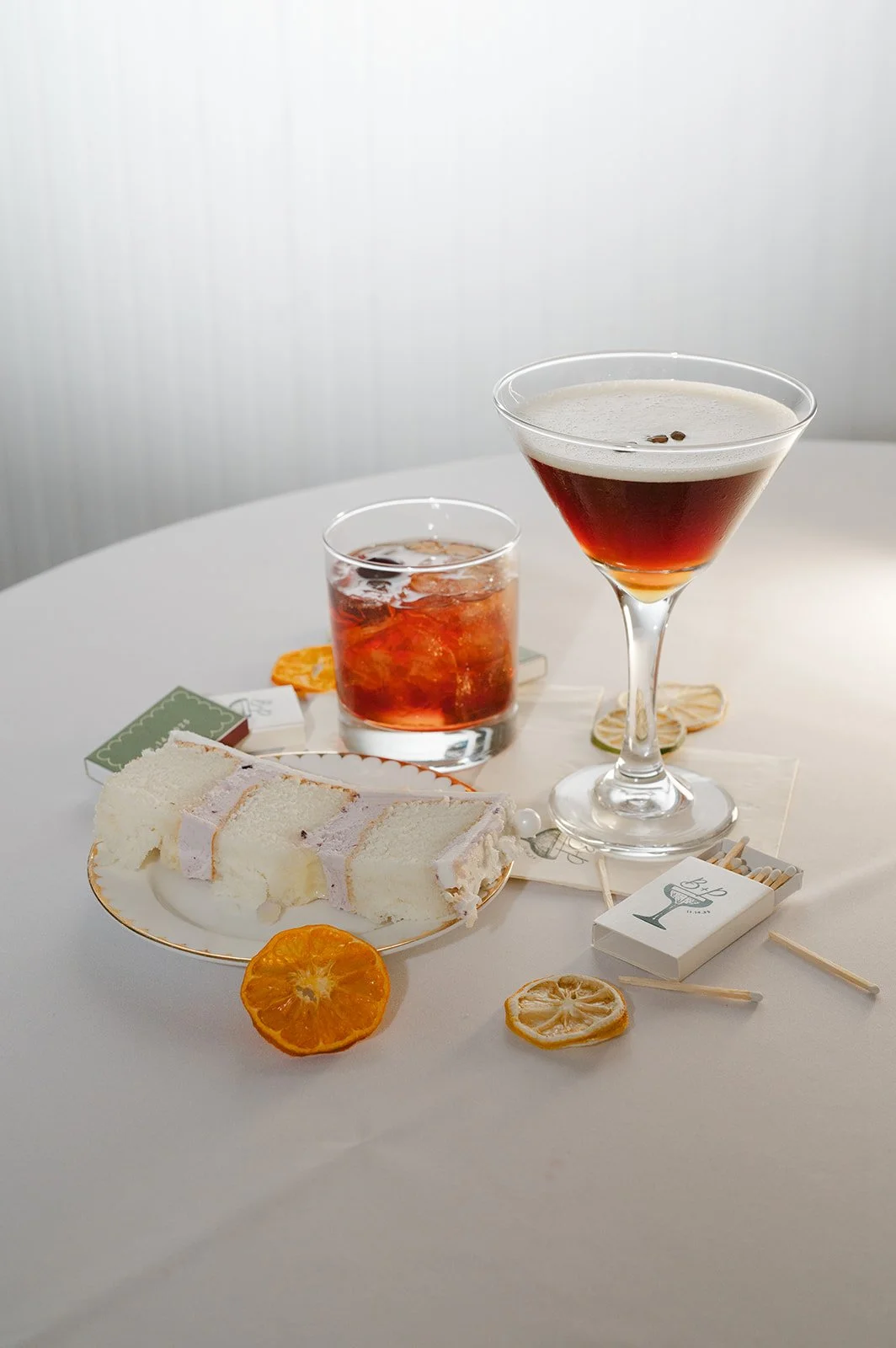 A white decorated cake on a gold stand surrounded by pink, white, and peach flowers on a wooden table, with gold cutlery and green napkins.